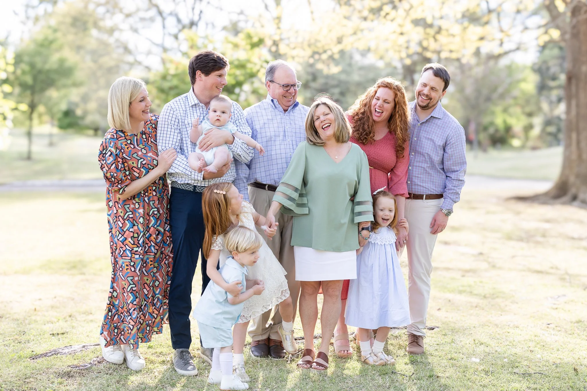 A family group photo in a park, including adults, children, and a baby, all smiling and wearing casual spring outfits.