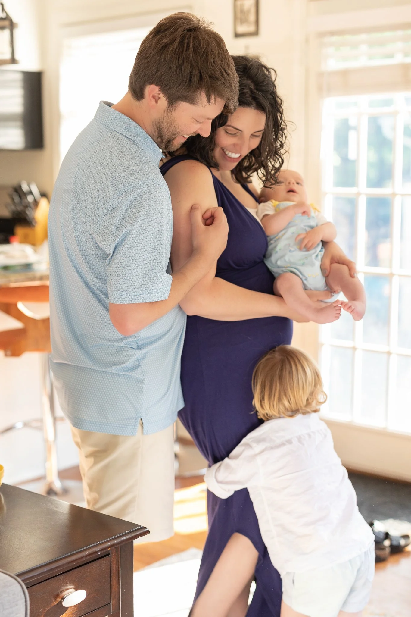 A family scene in a sunlit room with a man and woman smiling at each other, the woman holding a baby, and a young child hugging her leg.