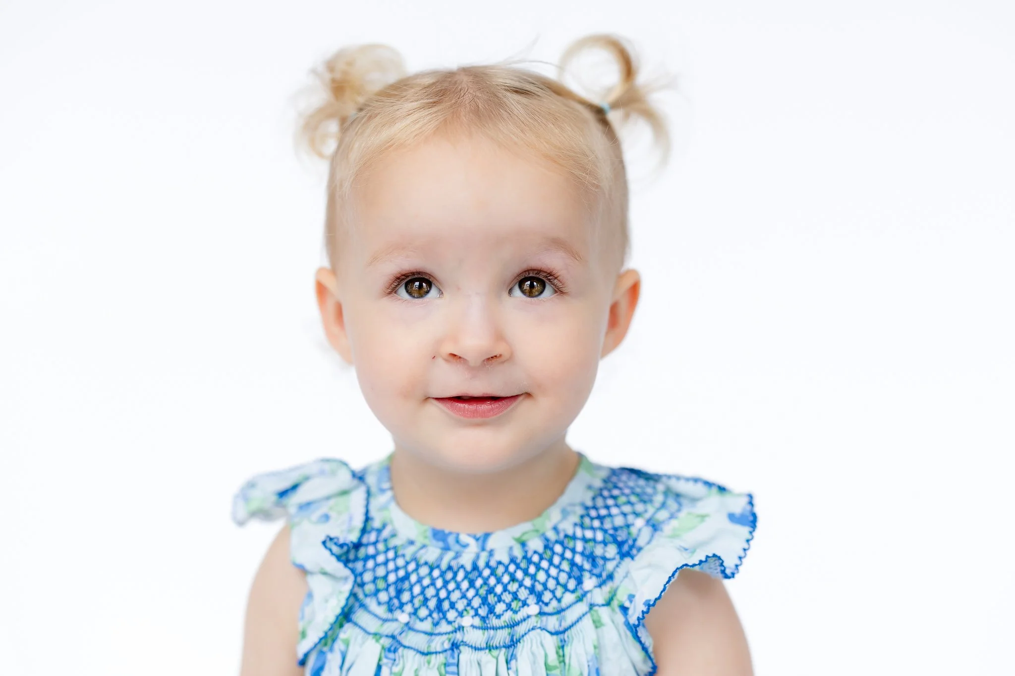 A toddler with blonde hair in pigtails wearing a blue and white patterned dress against a white background.