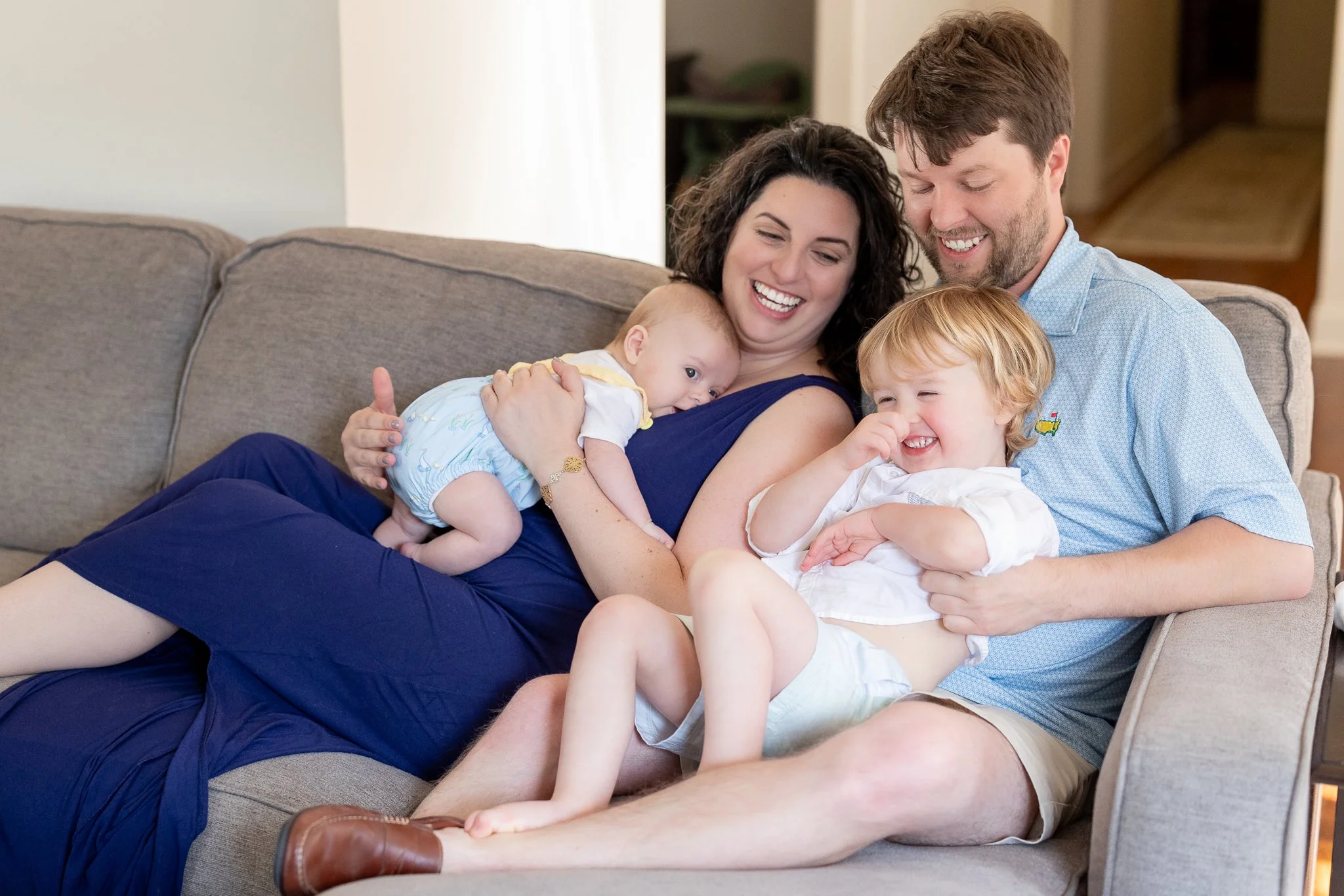 A happy family sitting on a couch, with a mother holding a baby and a father holding a toddler. The family is smiling and laughing, creating a joyful atmosphere.