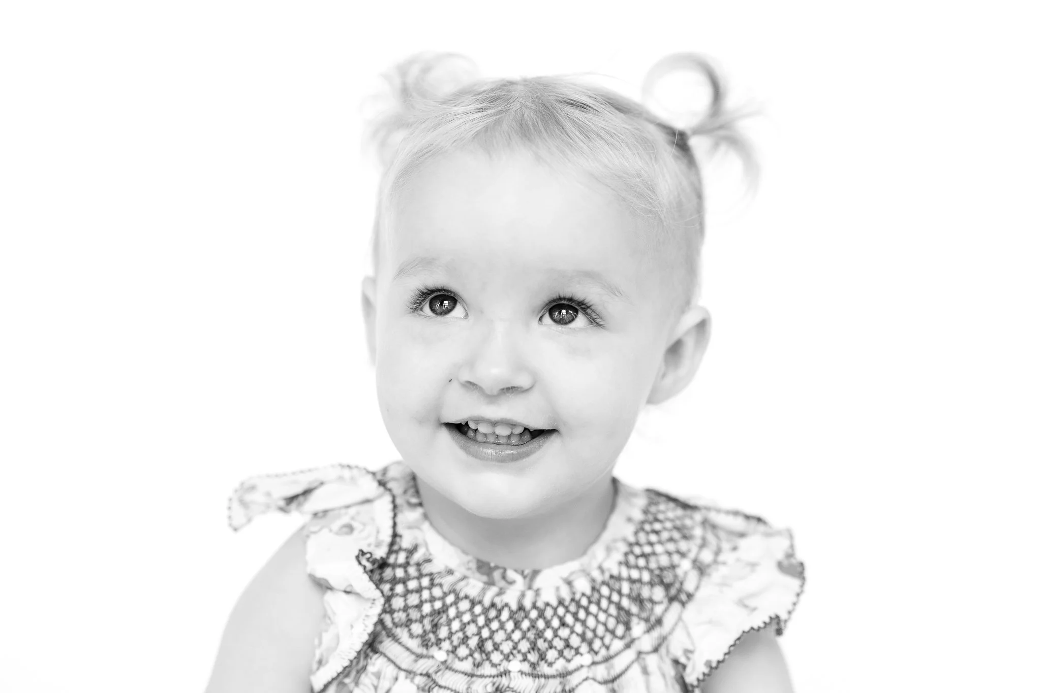 Baby smiling with curly hair in pigtails, wearing a patterned dress.