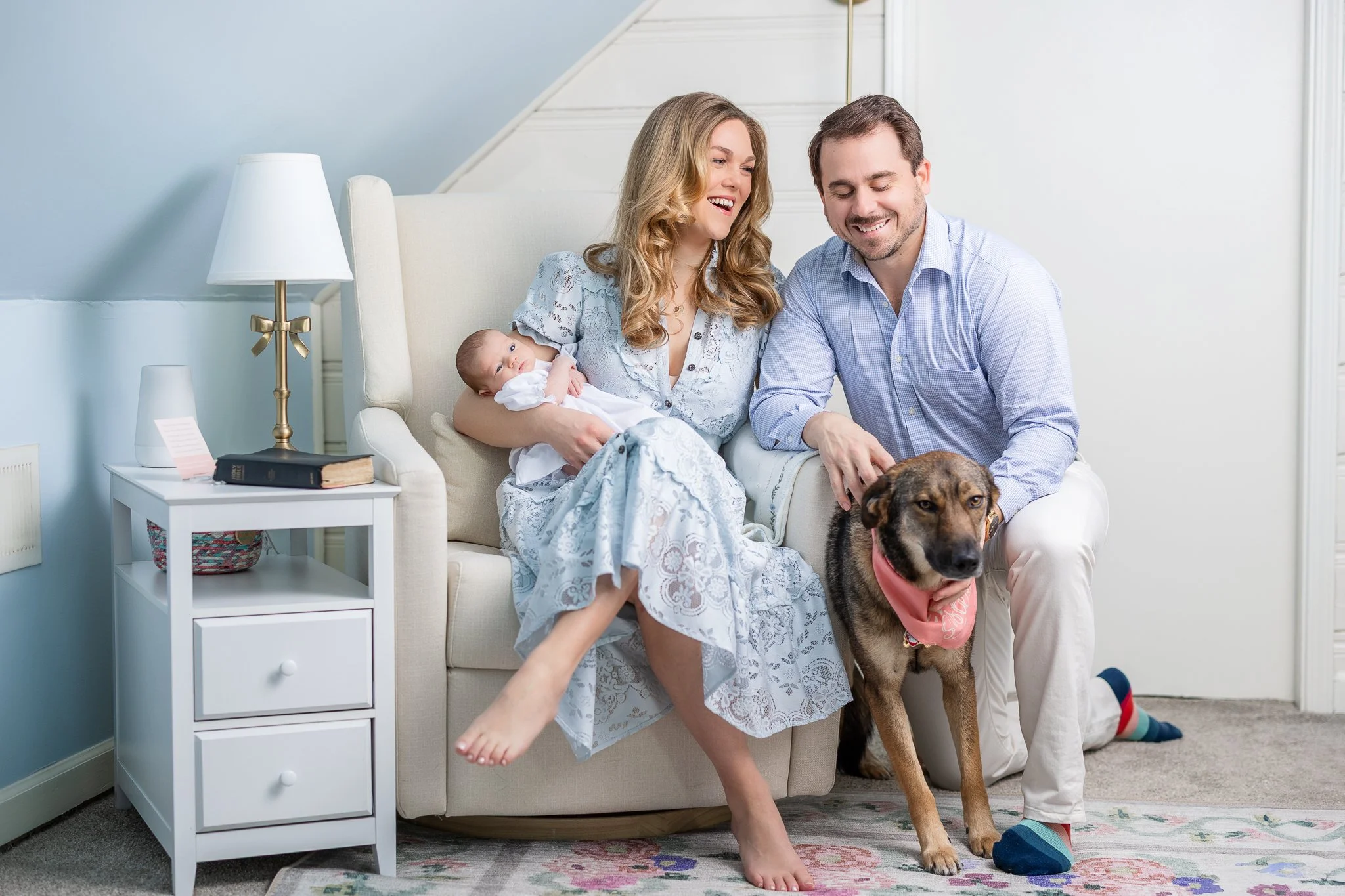 A couple smiling at home in a living room. The woman is holding a newborn baby, and the man is petting a dog wearing a bandana. They are sitting on a cream-colored chair next to a white side table with a lamp and a book. The room has a blue wall and 