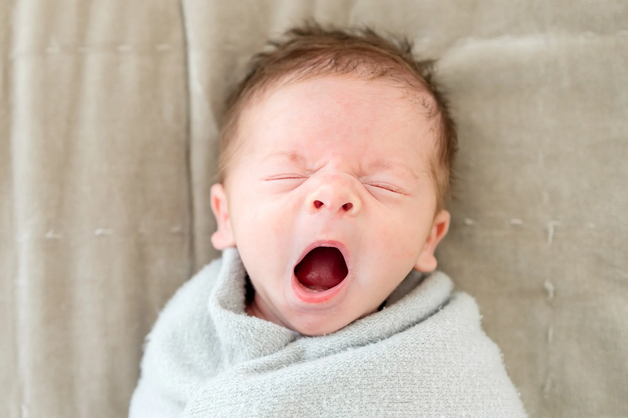 Yawning baby wrapped in a gray blanket on a soft surface.