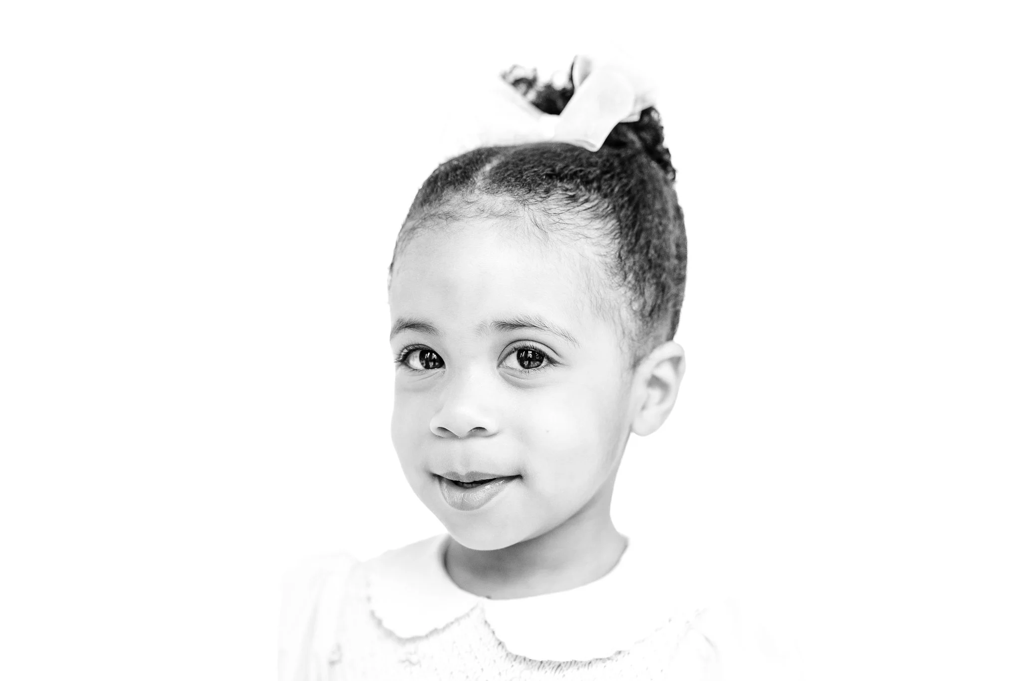 Black and white portrait of a young child with hair in a bun and a bow, wearing a collared dress, smiling slightly.