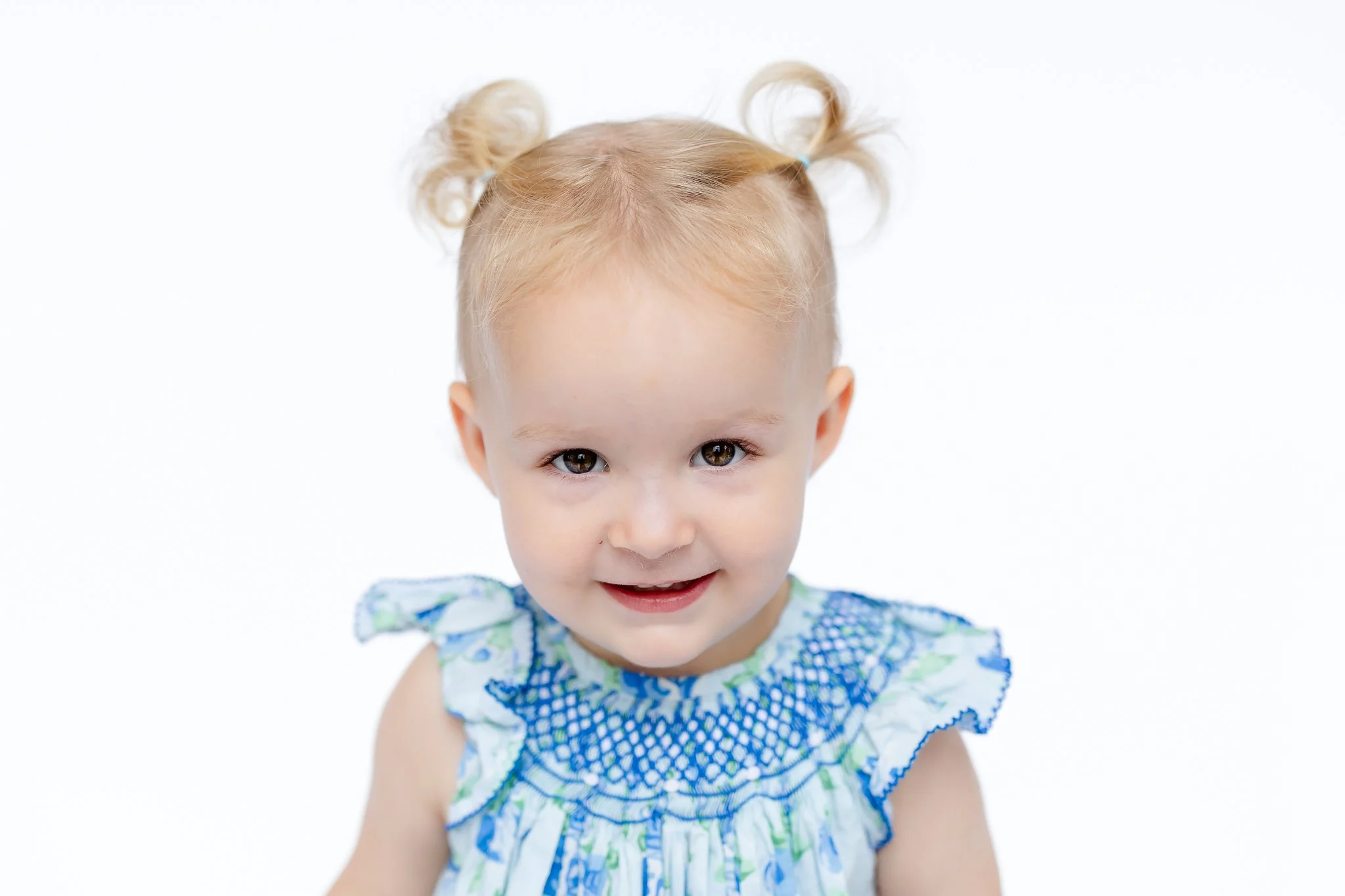 Toddler with blonde hair in pigtails, wearing a blue and green dress, smiling.