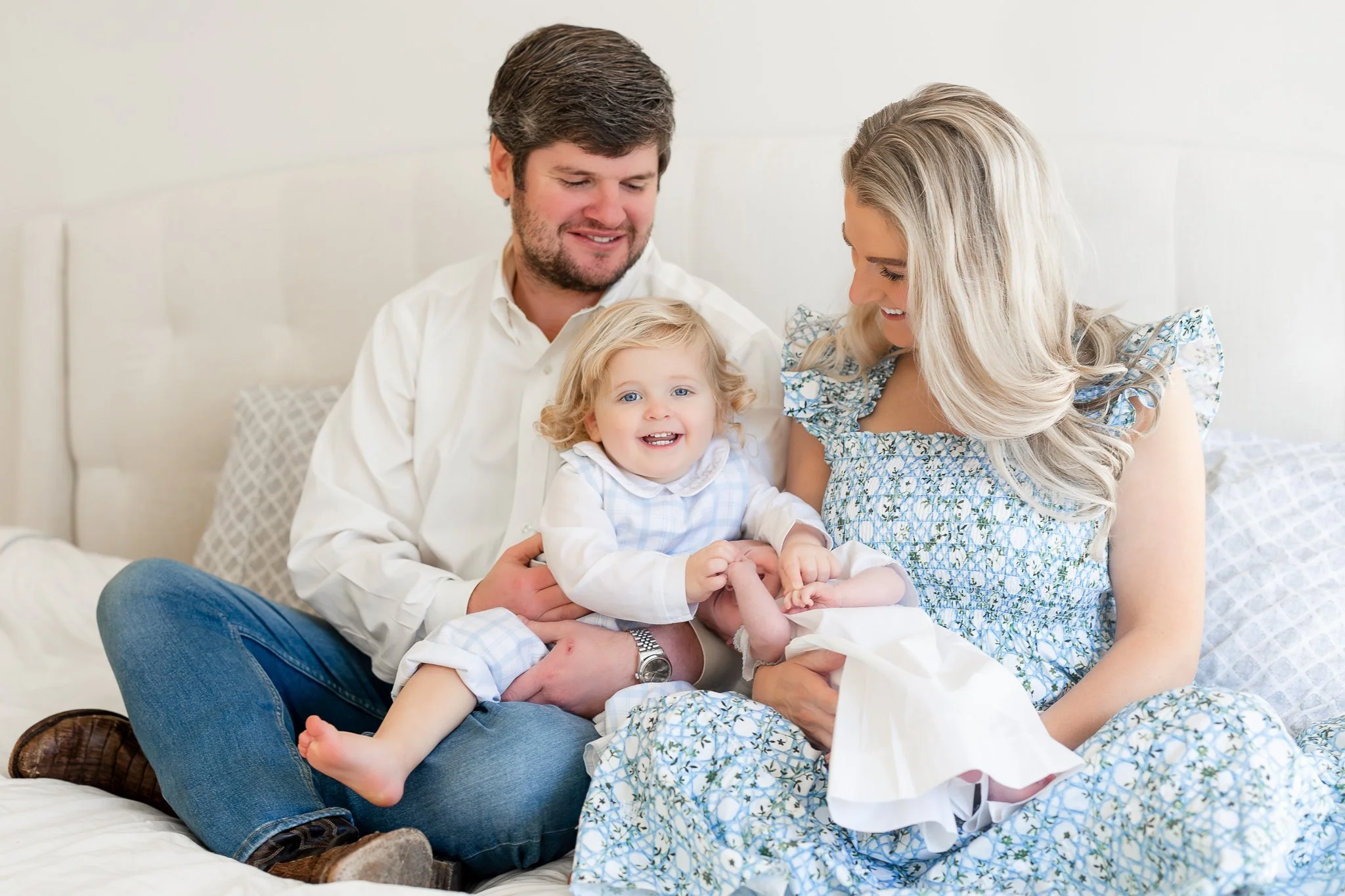 A family sitting on a bed with a smiling toddler and a baby. The man is wearing a white shirt, and the woman is wearing a blue floral dress. The toddler is in a light blue plaid outfit.
