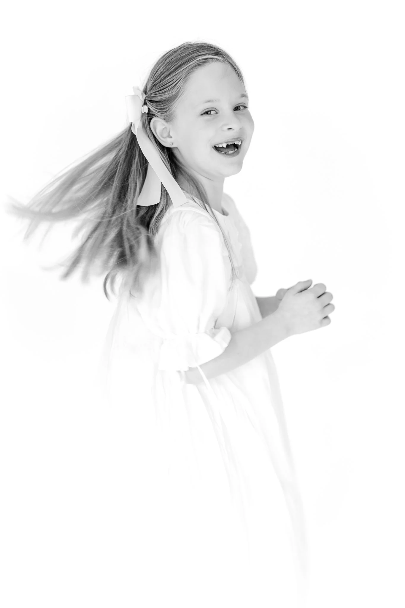 Black and white photo of a smiling girl in a white dress with a ribbon in her hair, looking to the side.