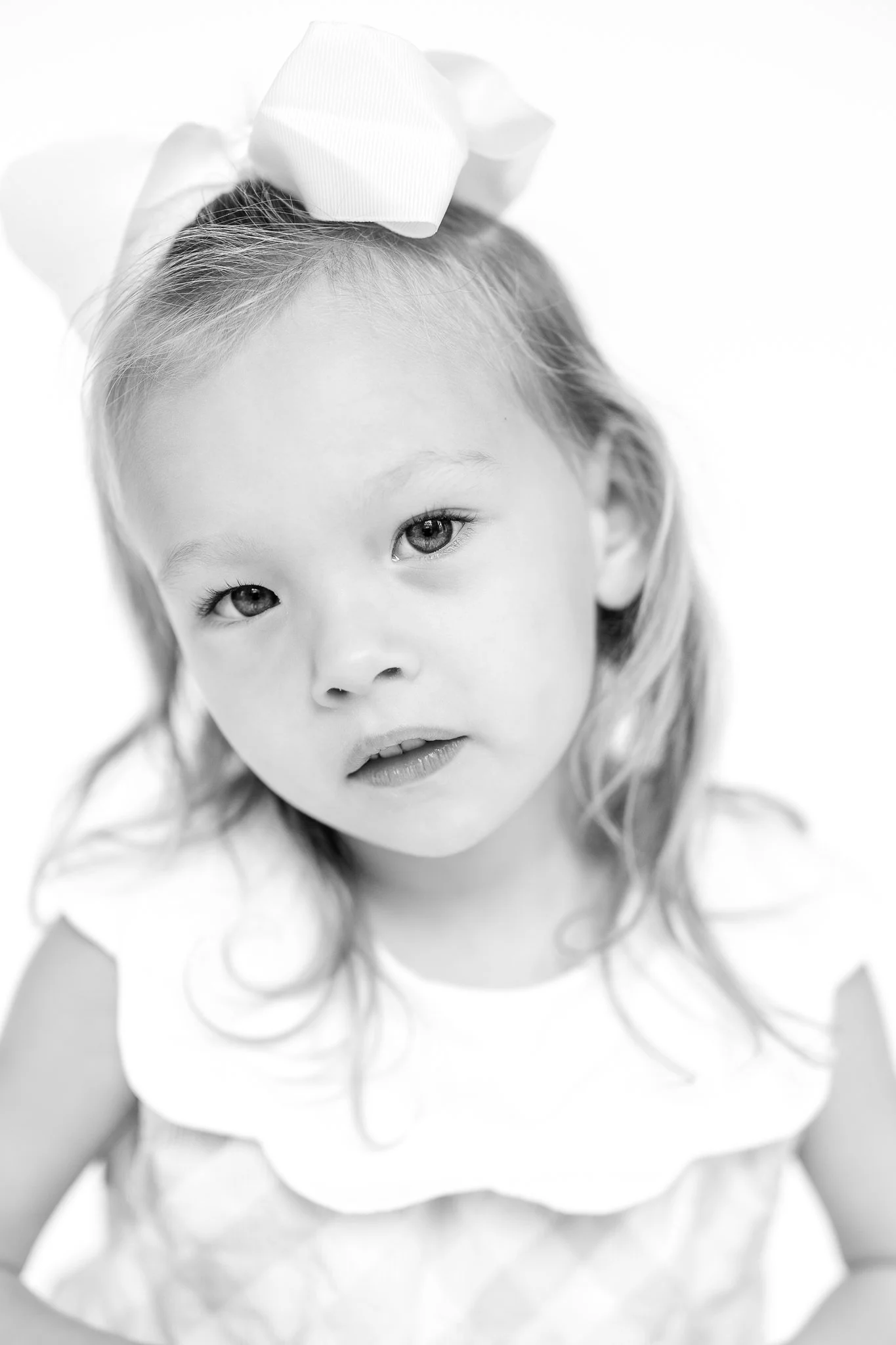 Black and white portrait of a young girl with a large hair bow, dressed in a patterned outfit, looking into the camera.