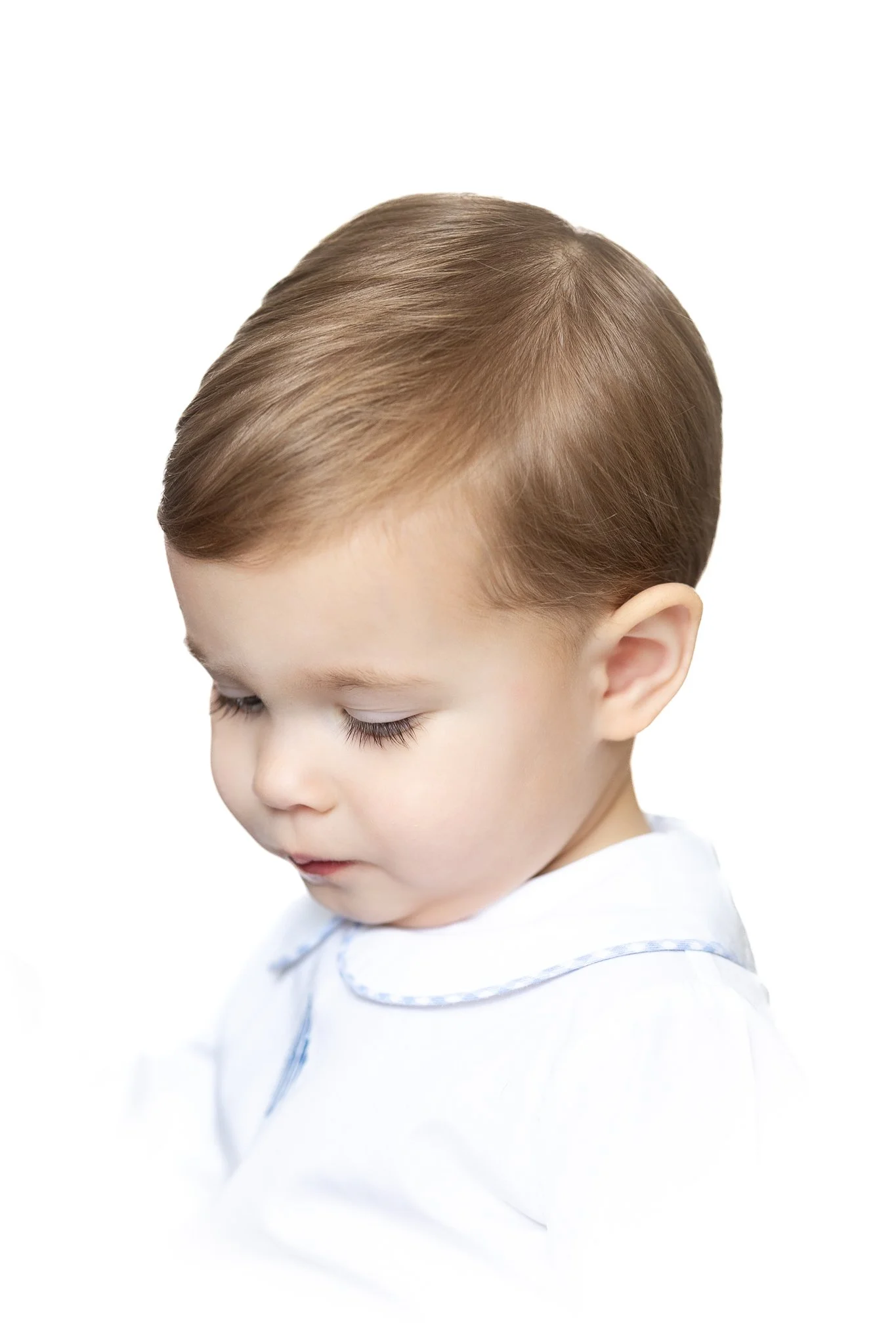 Profile view of a young child in a white outfit with light brown hair, looking downward against a white background.