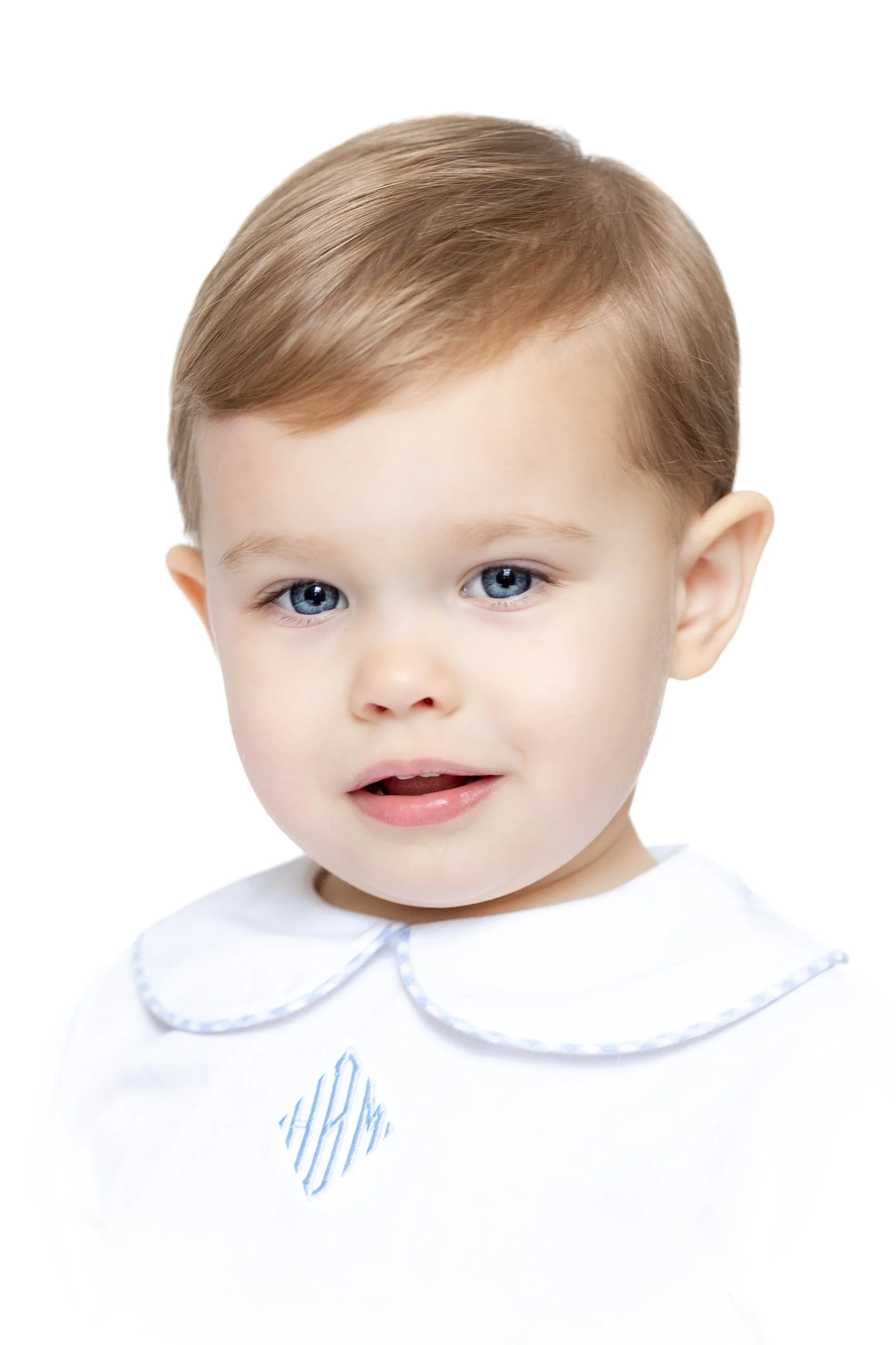 Toddler with short light brown hair, wearing a white shirt with blue trim and a decorative emblem, against a plain background in an heirloom portrait 