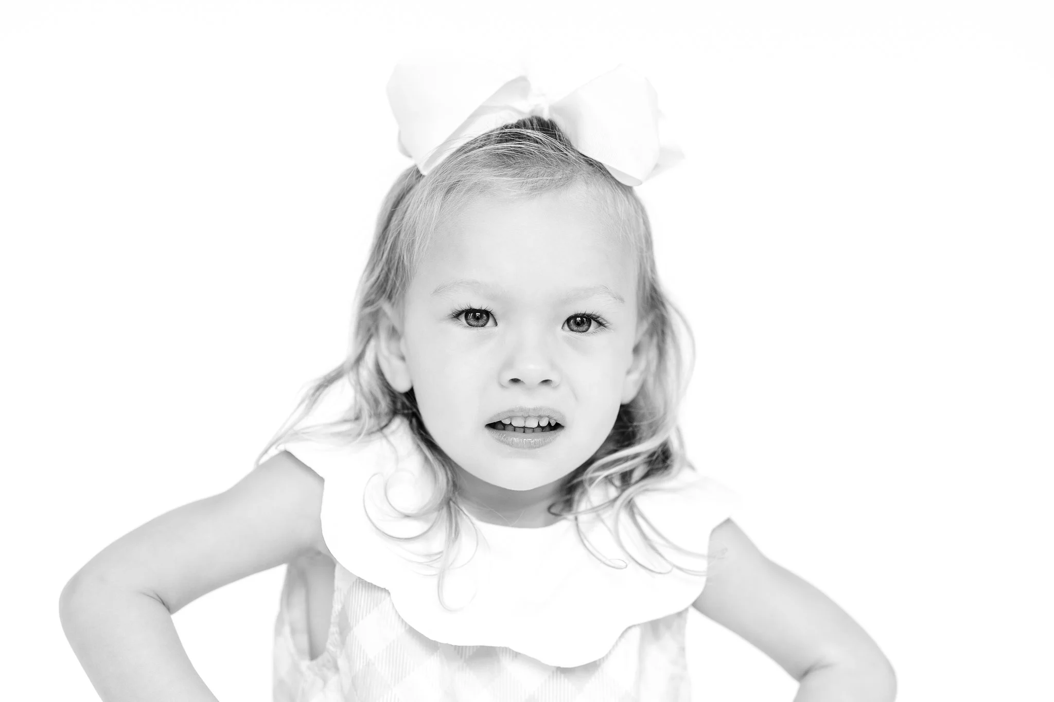 Black and white photo of a young girl with a large bow in her hair, looking at the camera, wearing a sleeveless dress with a scalloped collar.