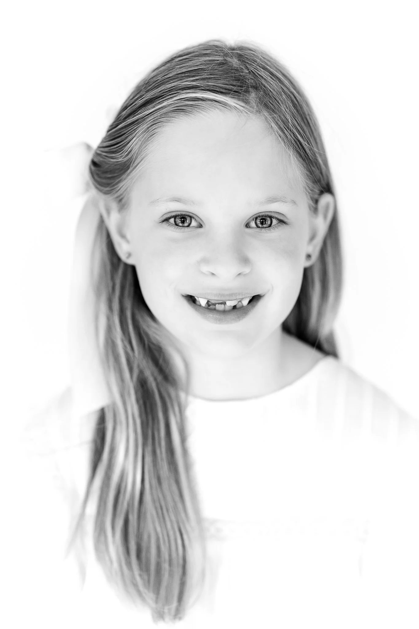 Black and white portrait of a smiling young girl with long hair, wearing a white top, and missing a front tooth.