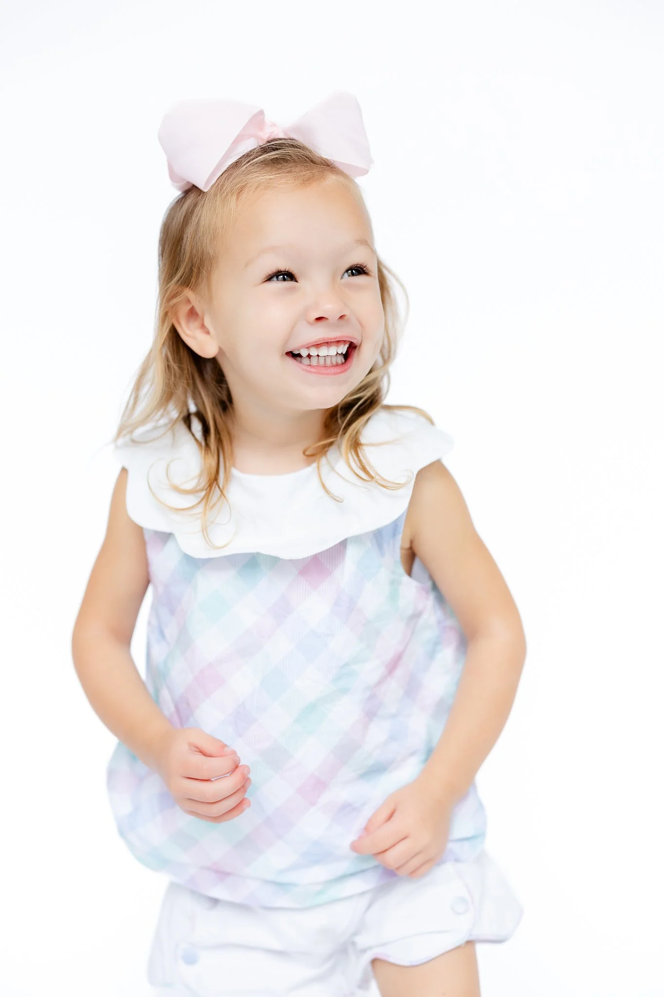 Happy child with a large pink bow, wearing a pastel plaid dress and white collar, smiling against a light background.