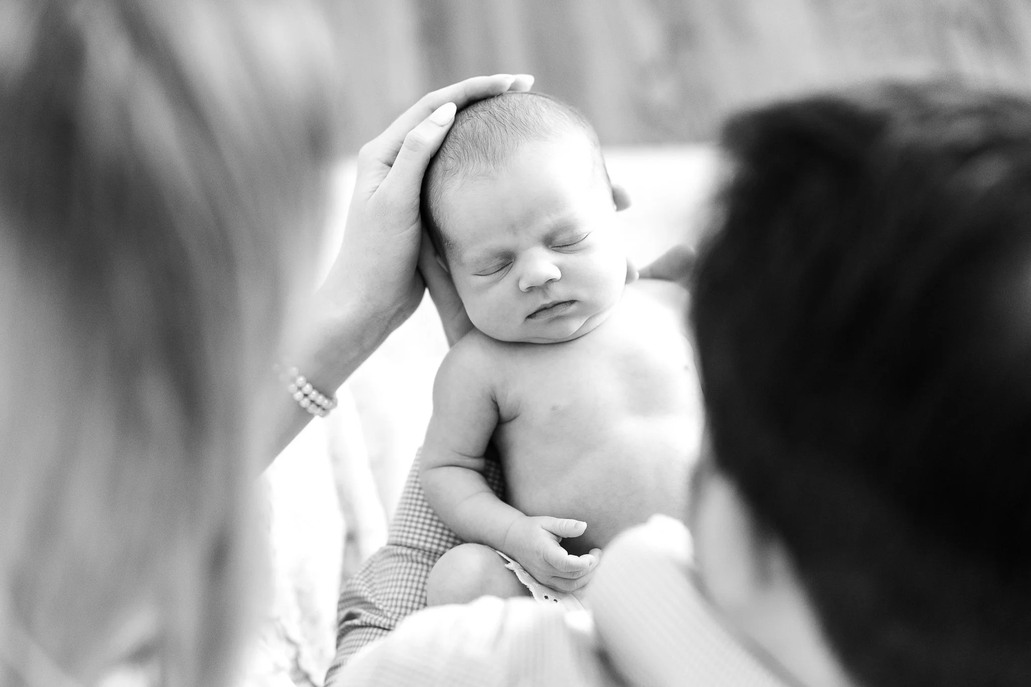 Black and white photo of a newborn baby being gently held and cradled by adults, with a hand resting on the baby's head.