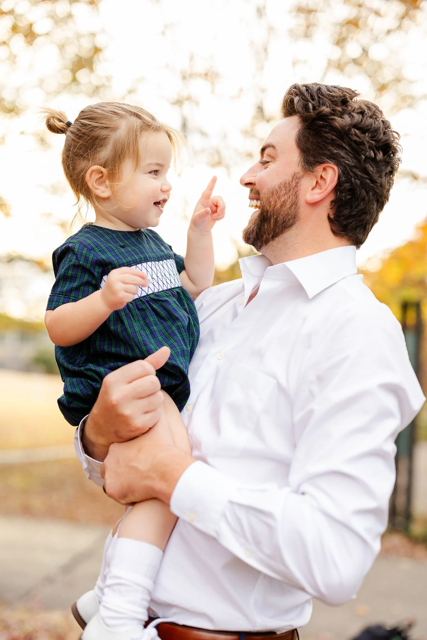 A smiling man holding a young child outdoors. The child is pointing at the man's face, with trees blurred in the background.