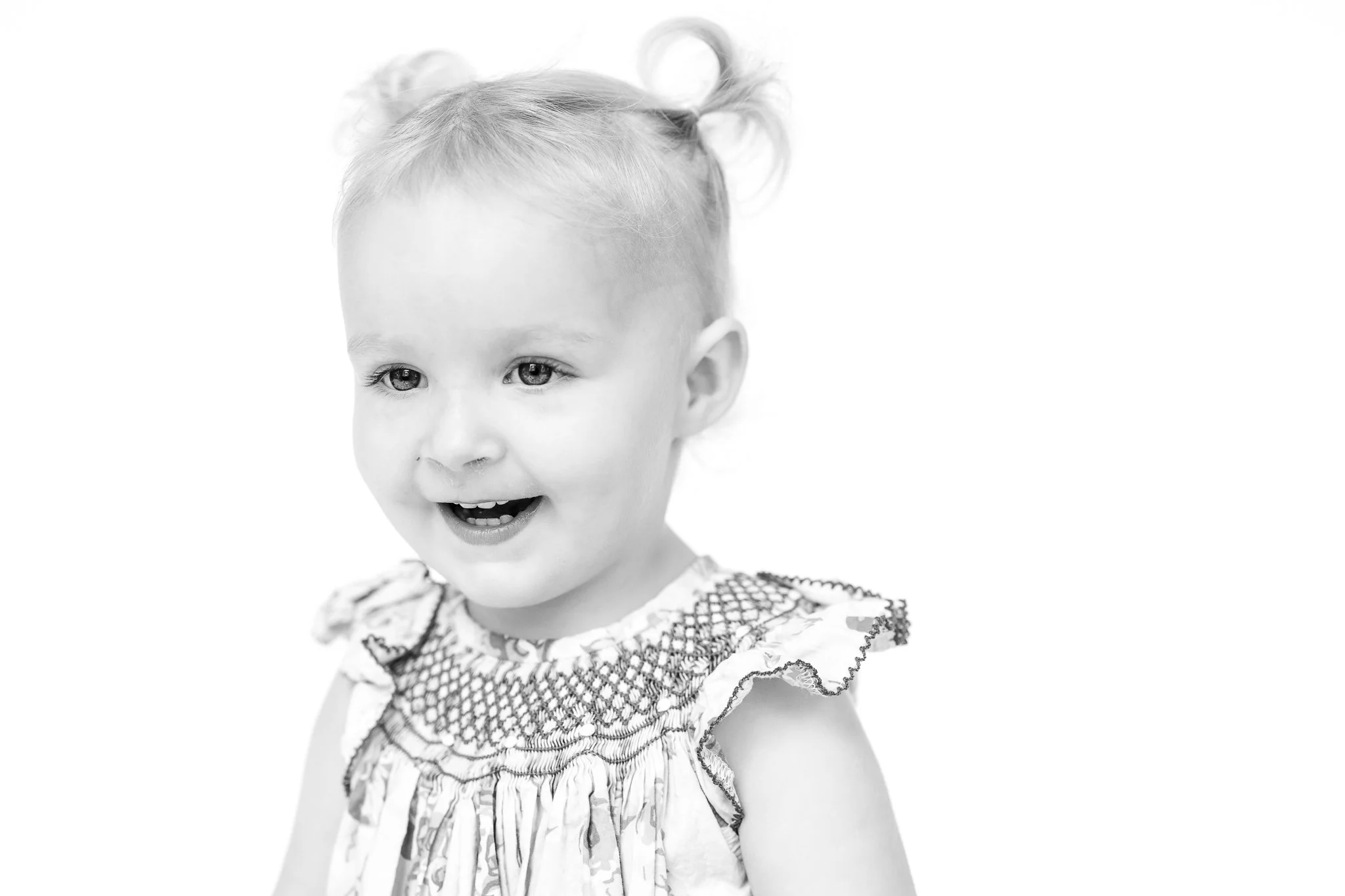 Black and white portrait of a smiling toddler with light hair and pigtails, wearing a patterned smocked dress.