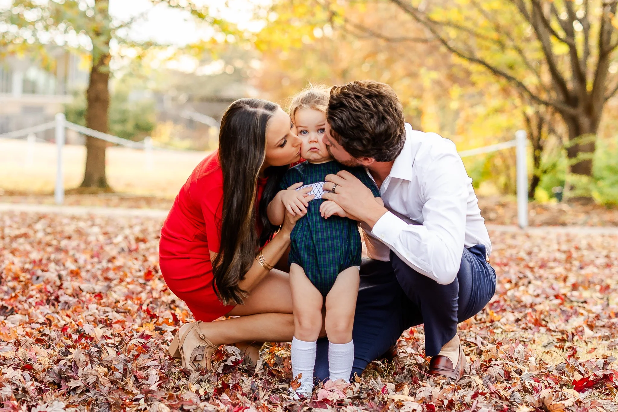 A family with a man, woman, and child posing in a park during fall, surrounded by autumn leaves. The man and woman are kissing the child's cheeks. The woman is wearing a red dress, and the man is in a white shirt and dark pants. The child is dressed 