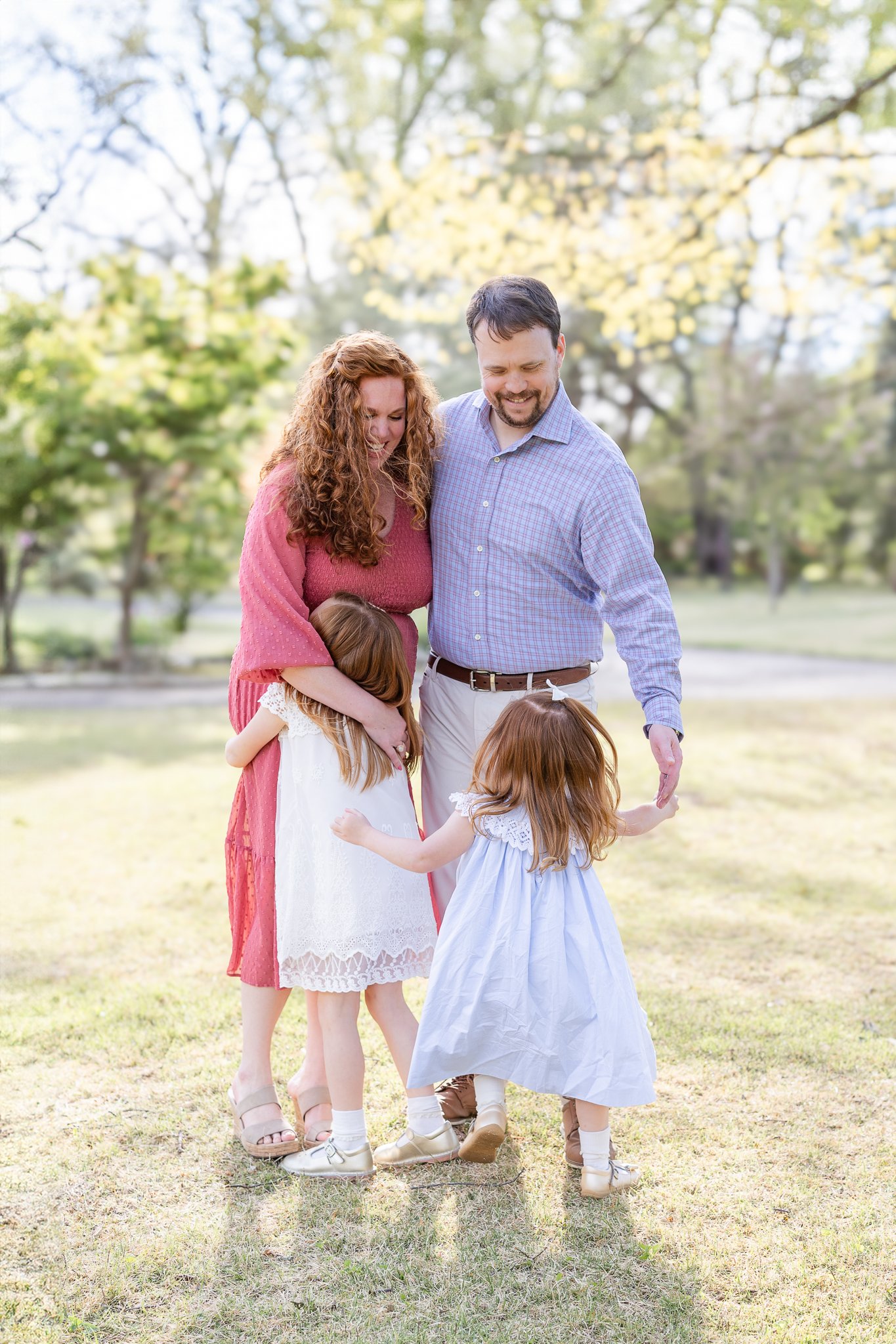 Family of four hugging in a park, with parents and two children, surrounded by trees and grass.