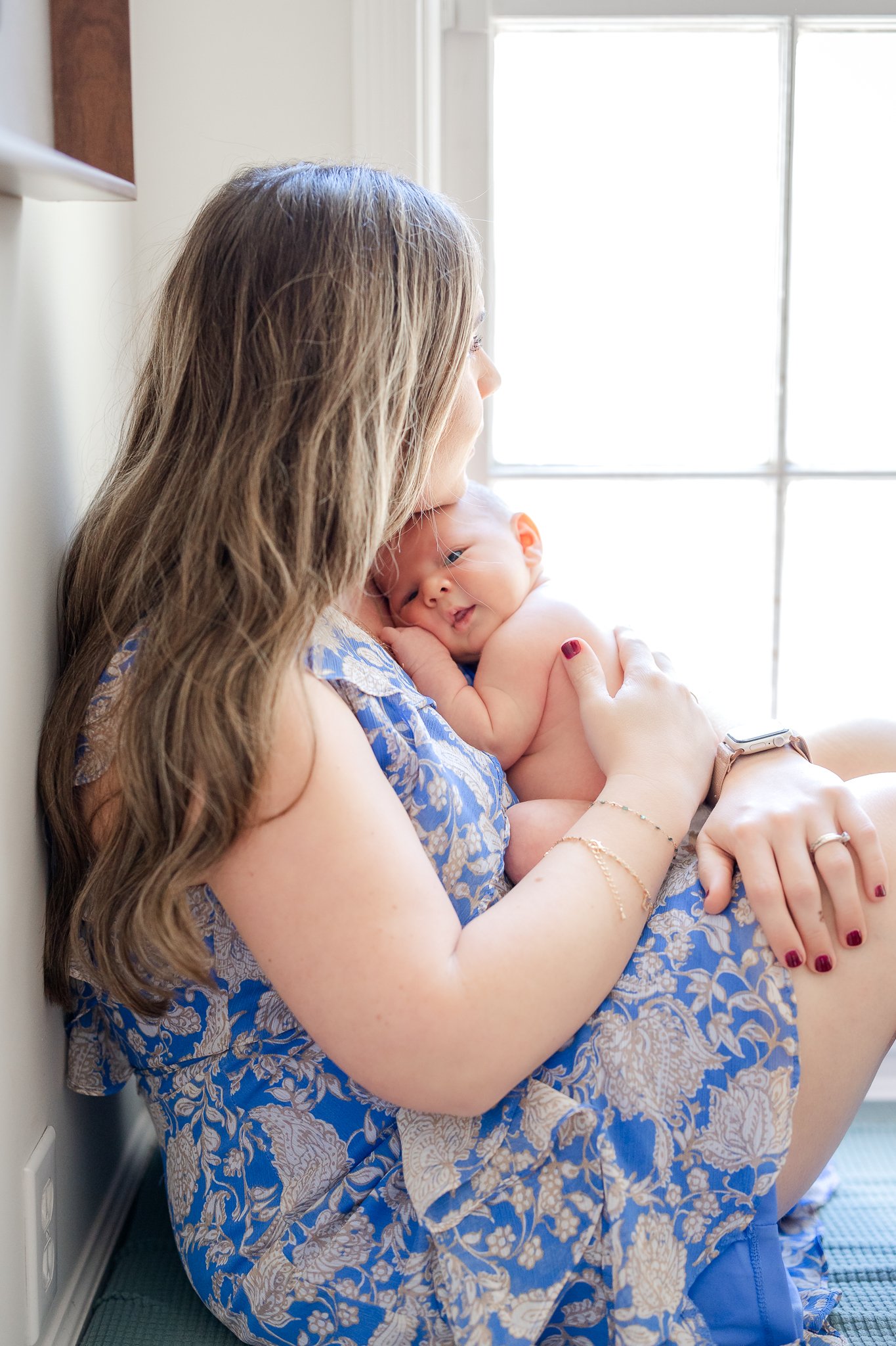 Woman in blue printed dress sitting by a window, holding a baby close to her chest.