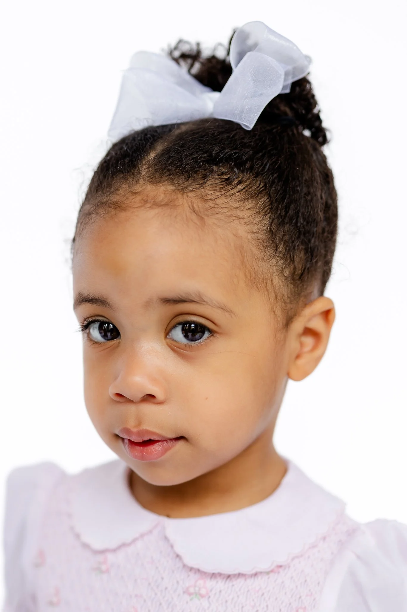 Close-up of a young child with curly hair, wearing a large bow and a white dress with a collar.