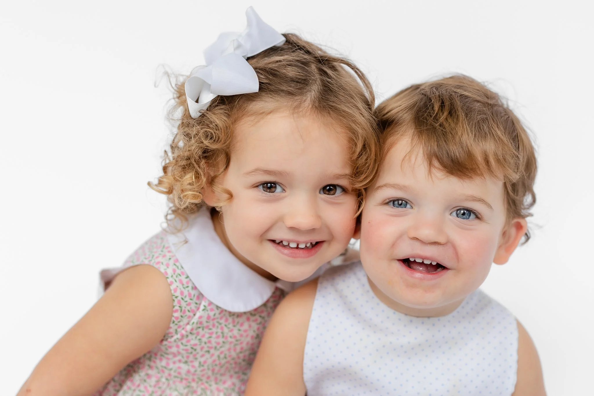 Two young children smiling, one with curly hair wearing a floral dress and hair bow, the other wearing a polka dot top.