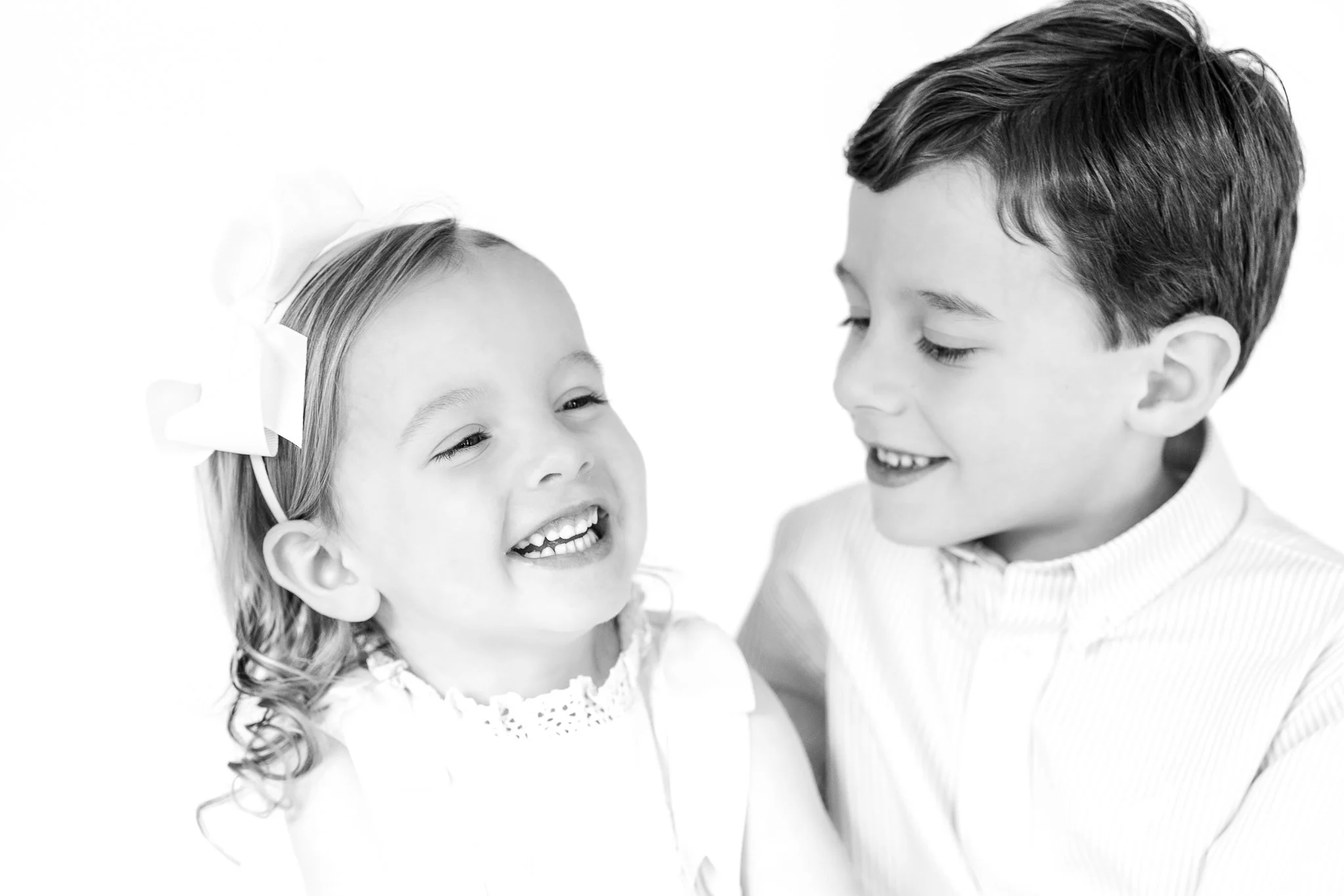 Black and white portrait of two smiling children, a girl with a bow in her hair and a boy looking at her.