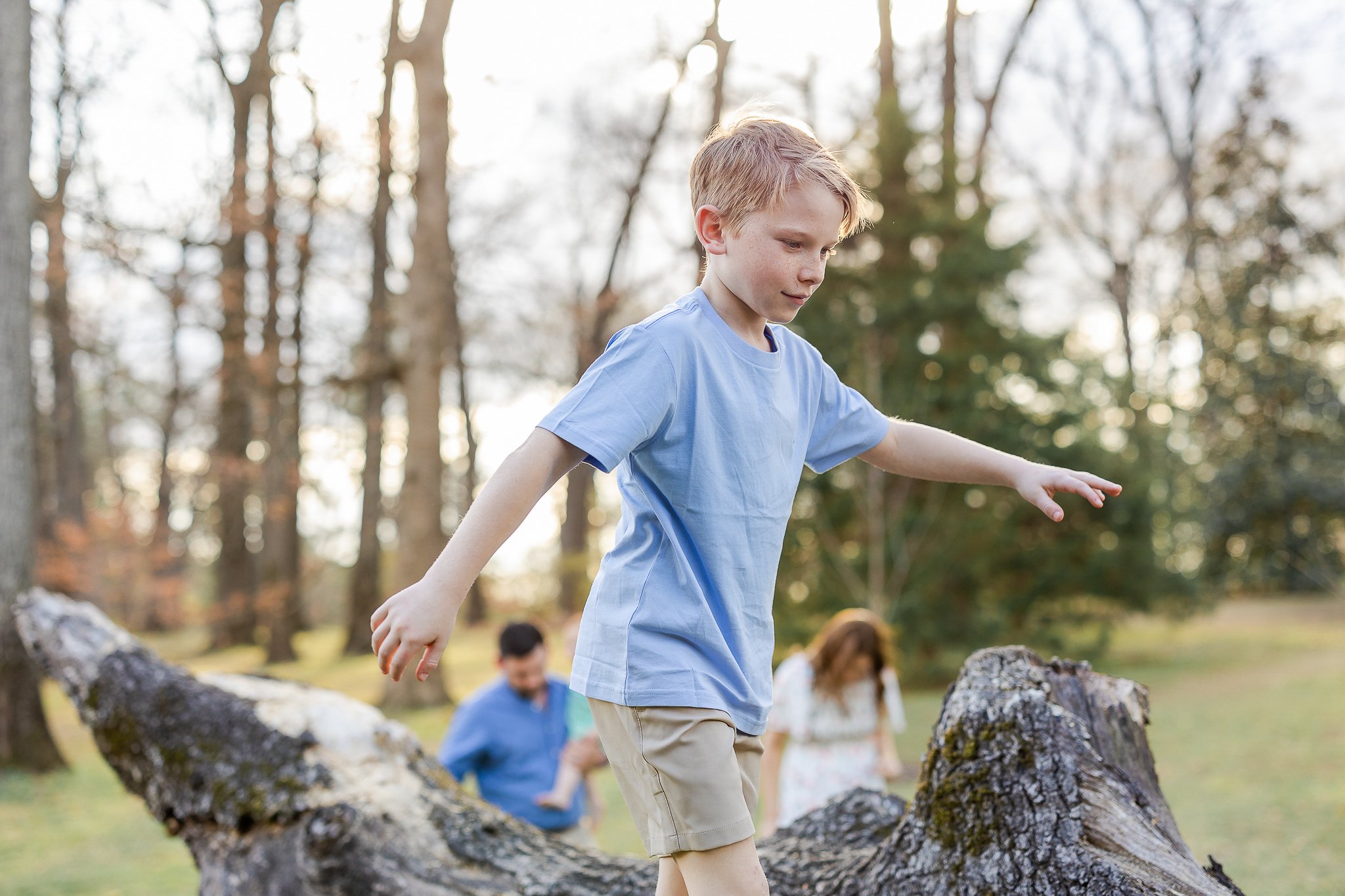 A young boy in a light blue T-shirt balancing on a fallen tree in a forest, with blurred adults in the background.