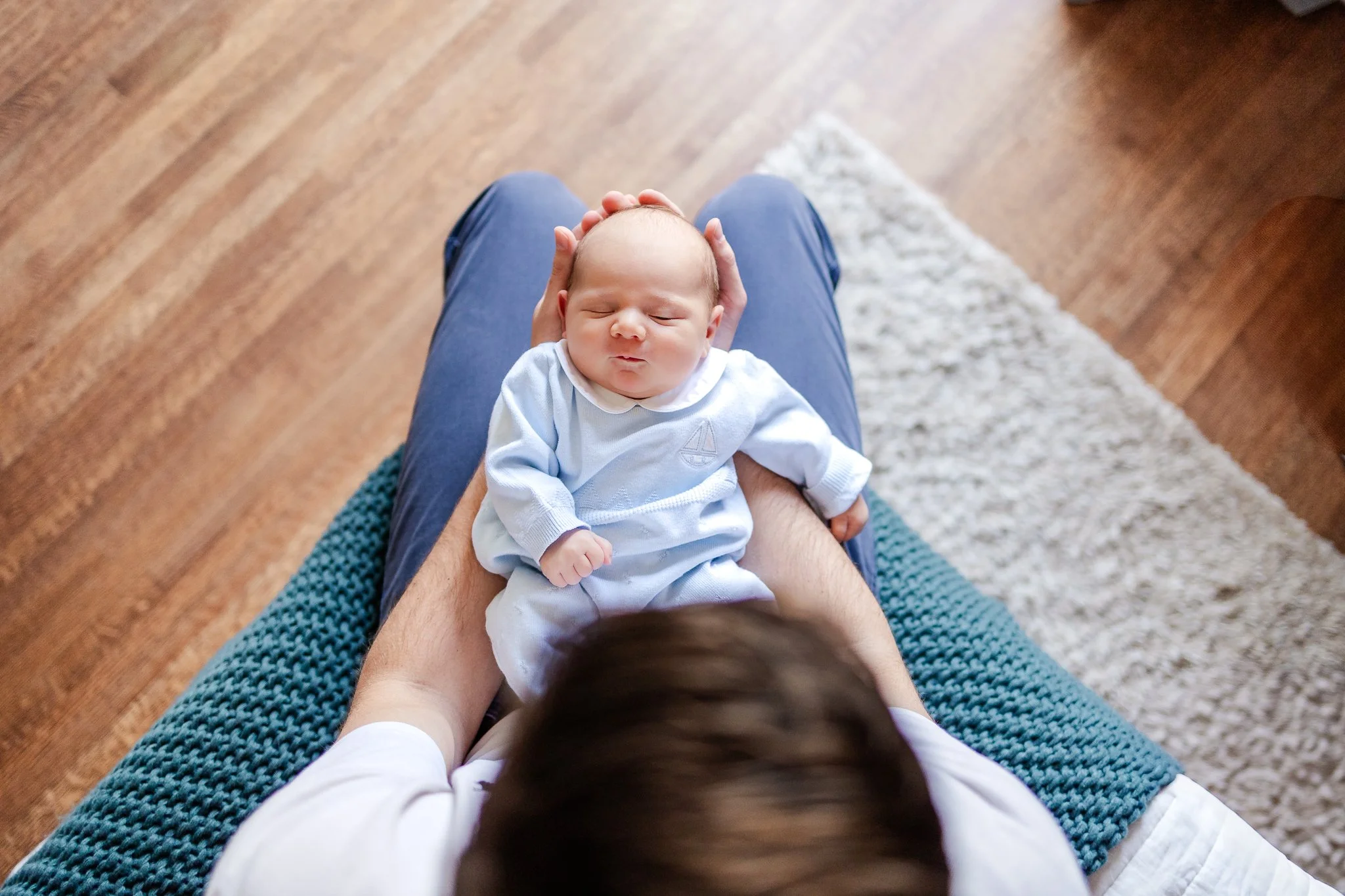 Person holding a newborn baby on their lap on a wooden floor with a rug nearby.