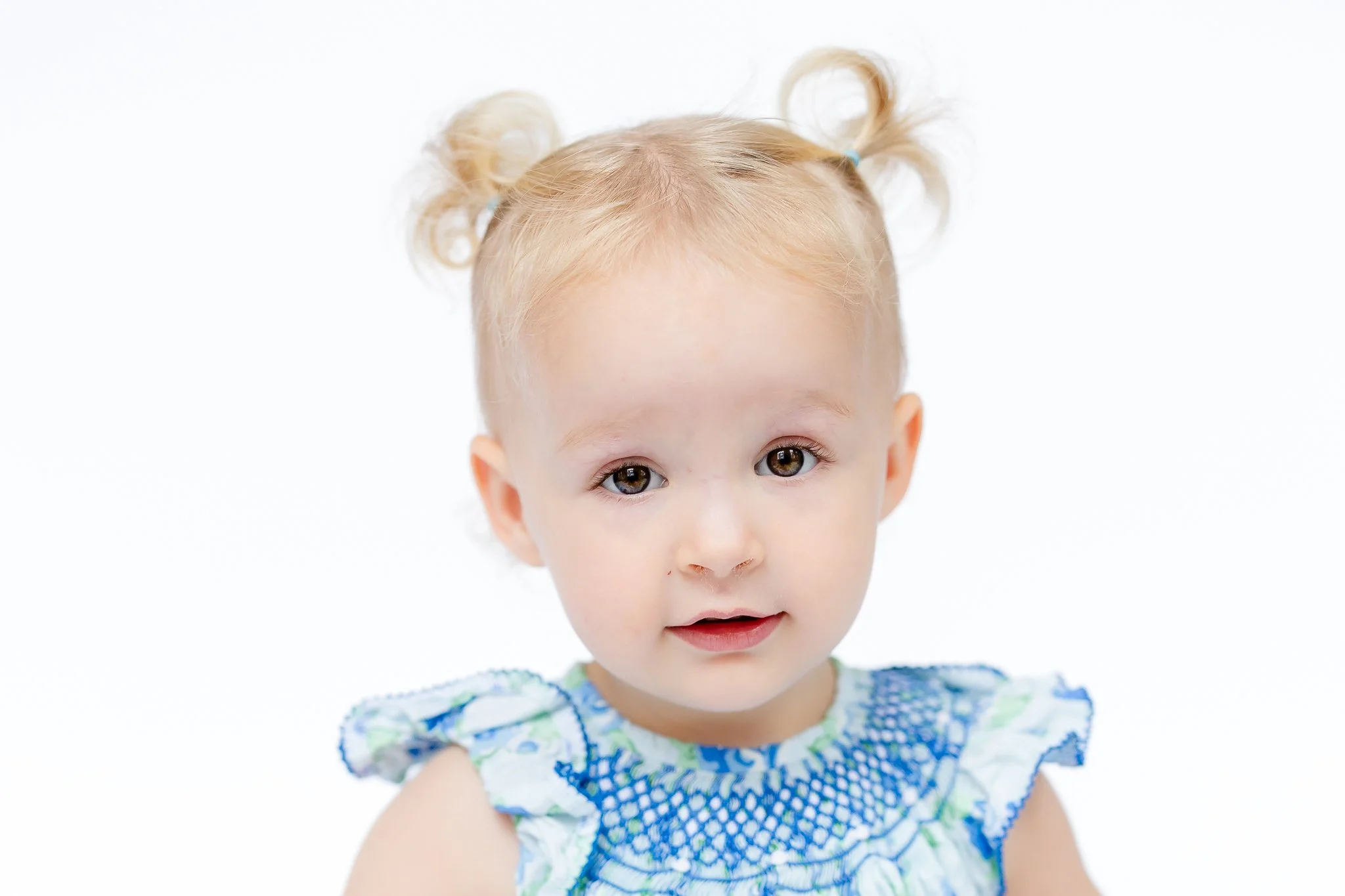 Baby with blonde hair in pigtails wearing a blue and white dress, against a white background.