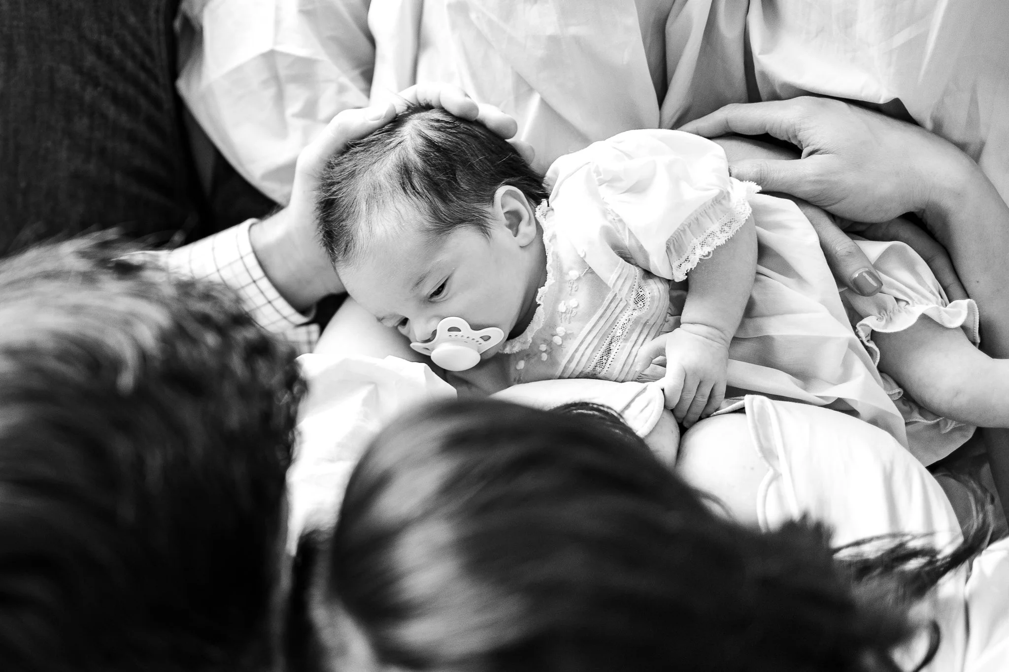 Black and white image of a newborn baby with a pacifier, lying on an adult's lap, surrounded by family members.