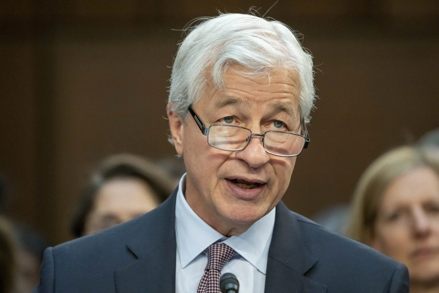 An older man with white hair and glasses speaking in a formal setting, wearing a navy suit, white shirt, and patterned tie, with a microphone in front of him.