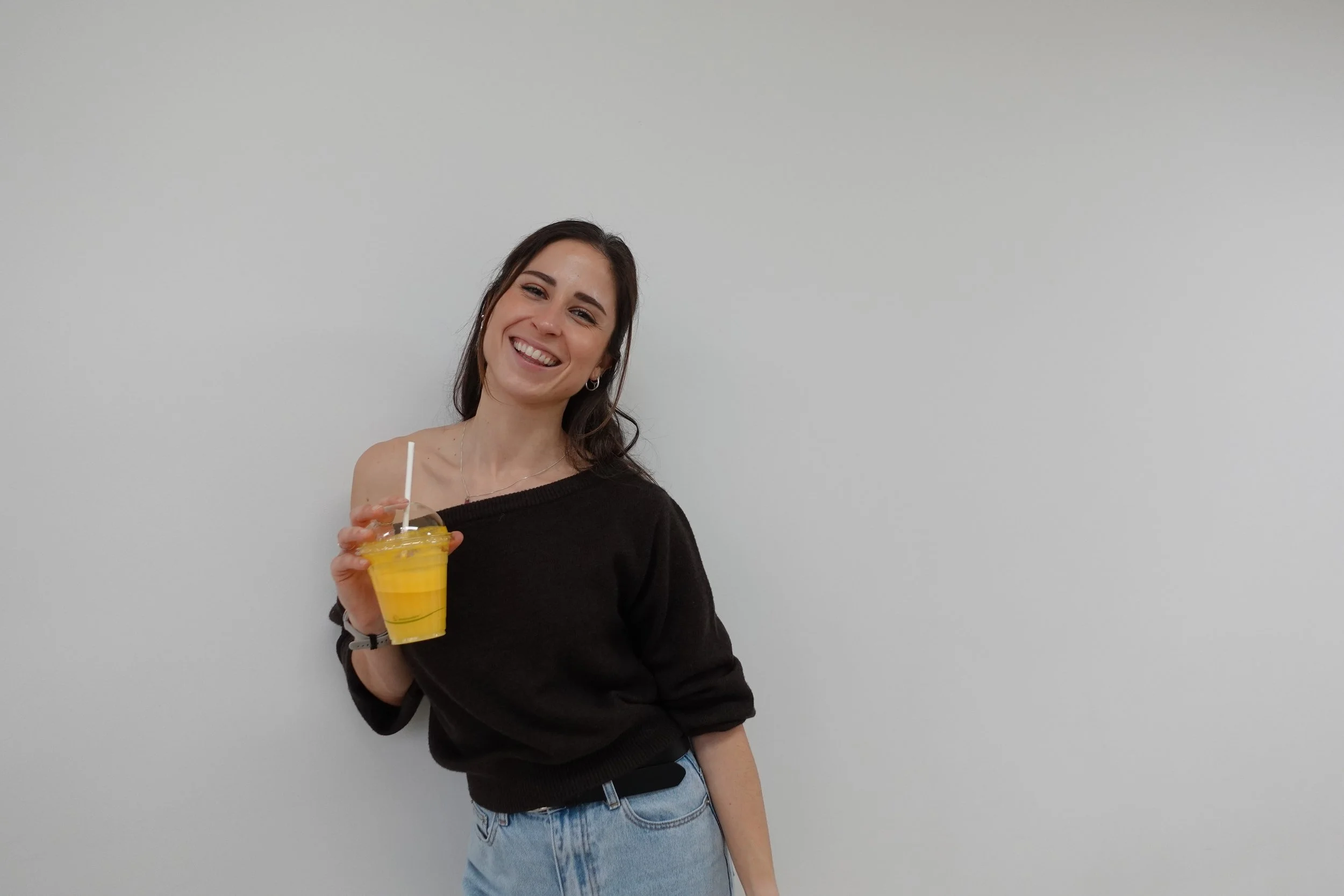 Mon Kinloch smiling wearing a brown off the shoulder jumper, holding a drink of orange juice in her hand