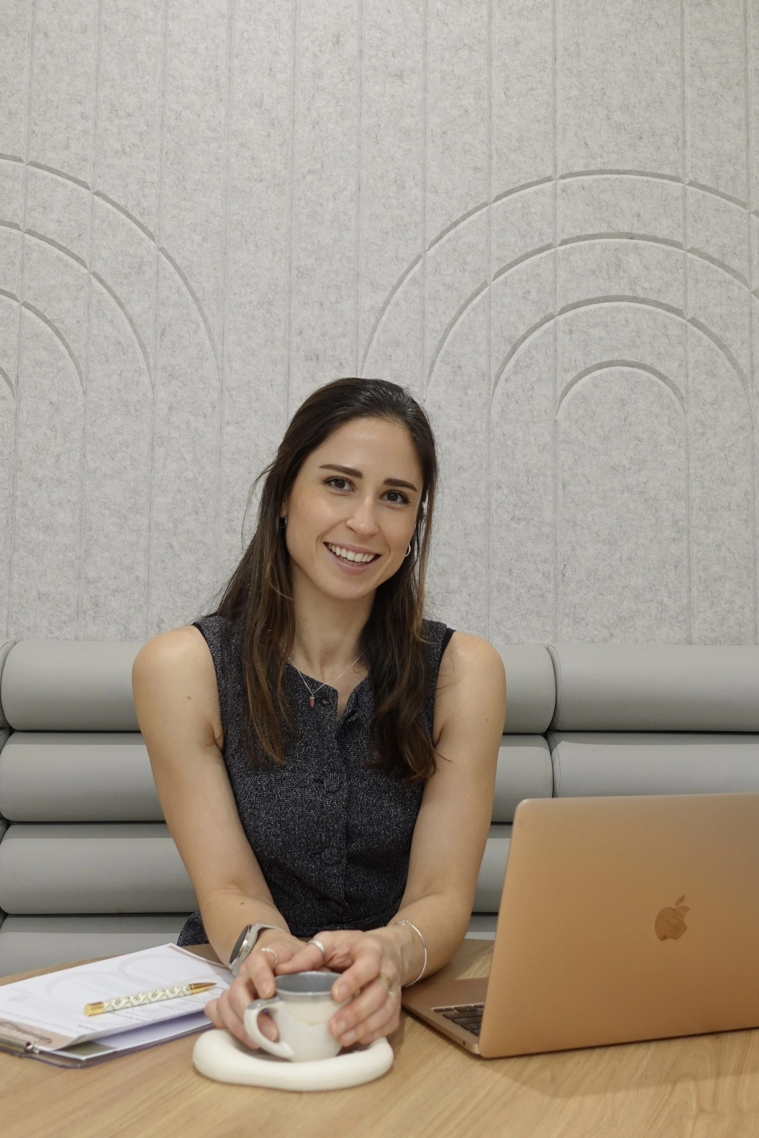 Mon sitting casually at a desk wearing a grey sleeveless work top, smiling at the camera and holding a tea cup in her hands, between a note pad and pen and a laptop
