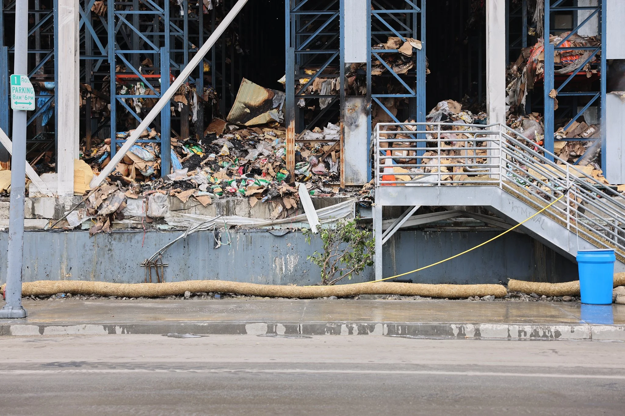 Trapped between charred, towering, collapsed racks, cardboard and plastic storage boxes are crushed by the massive weight of the roof and the water used to battle the blaze on July 2, 2026, in Los Angeles, CA. 