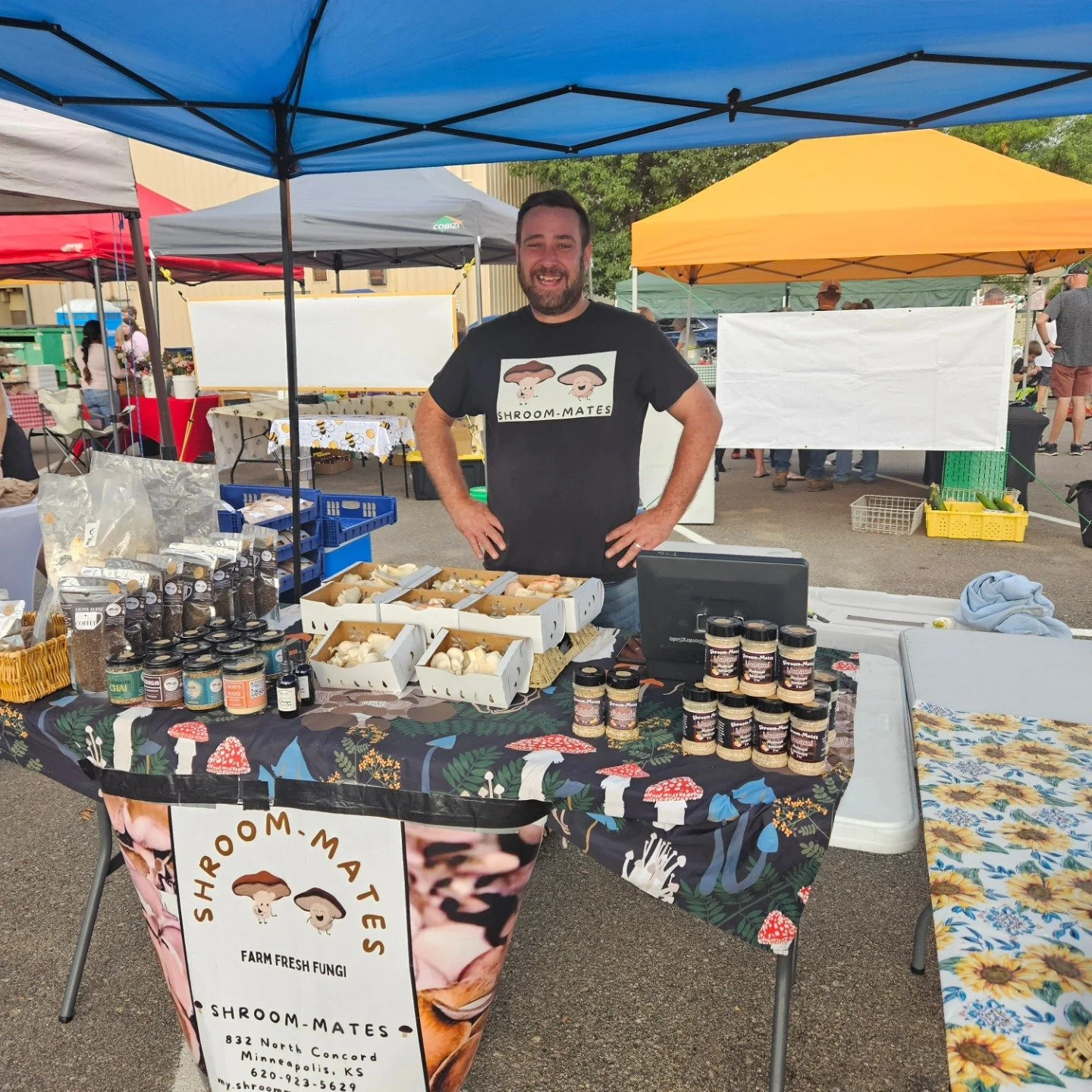 A man standing behind a table with farm fresh fungi at a farmers market booth. The table displays boxes of mushrooms and a sign with the stall's name and contact information.