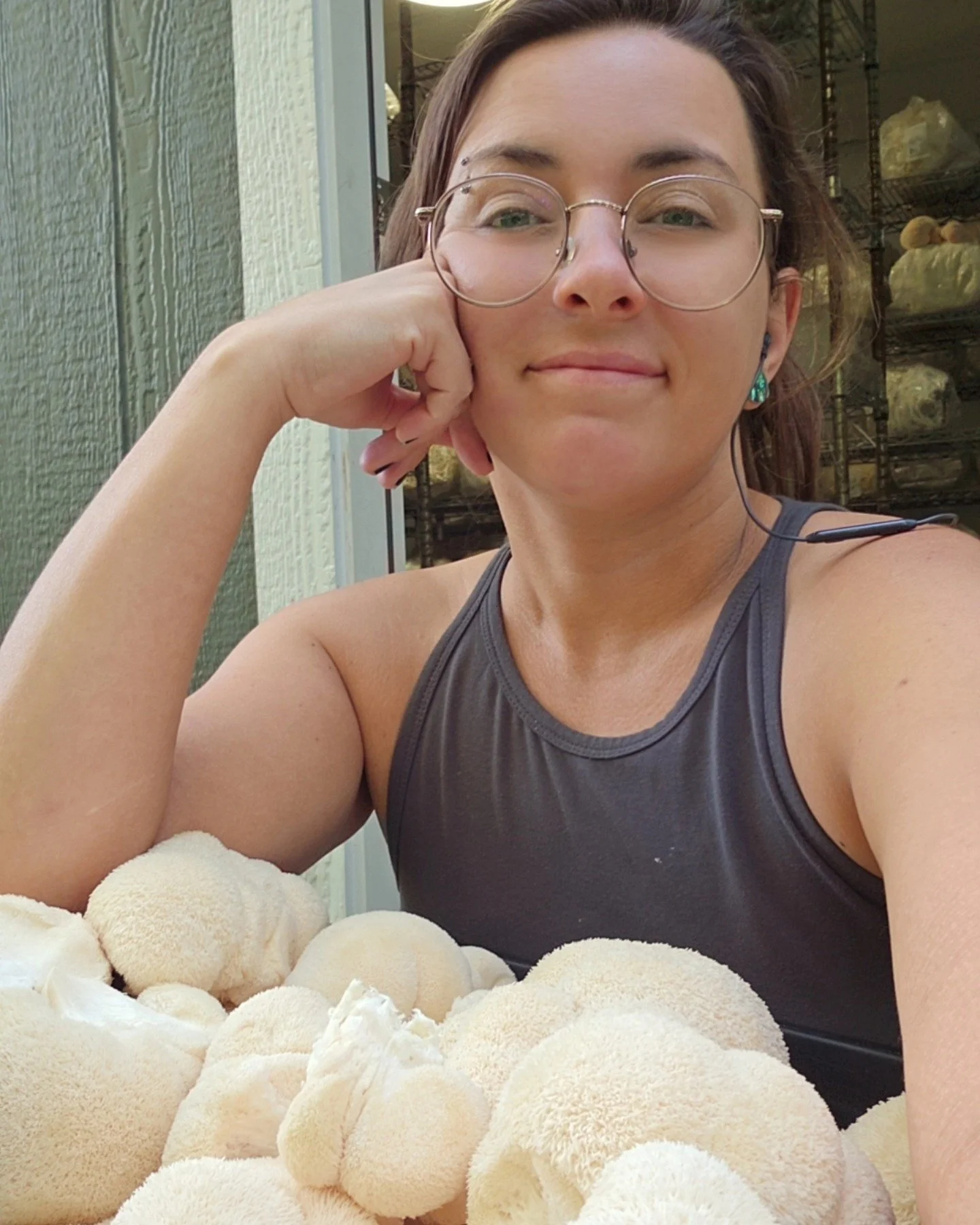 A woman in a gray t-shirt, glasses, and a baseball cap holding two boxes of mushrooms inside a mushroom farm or shop.