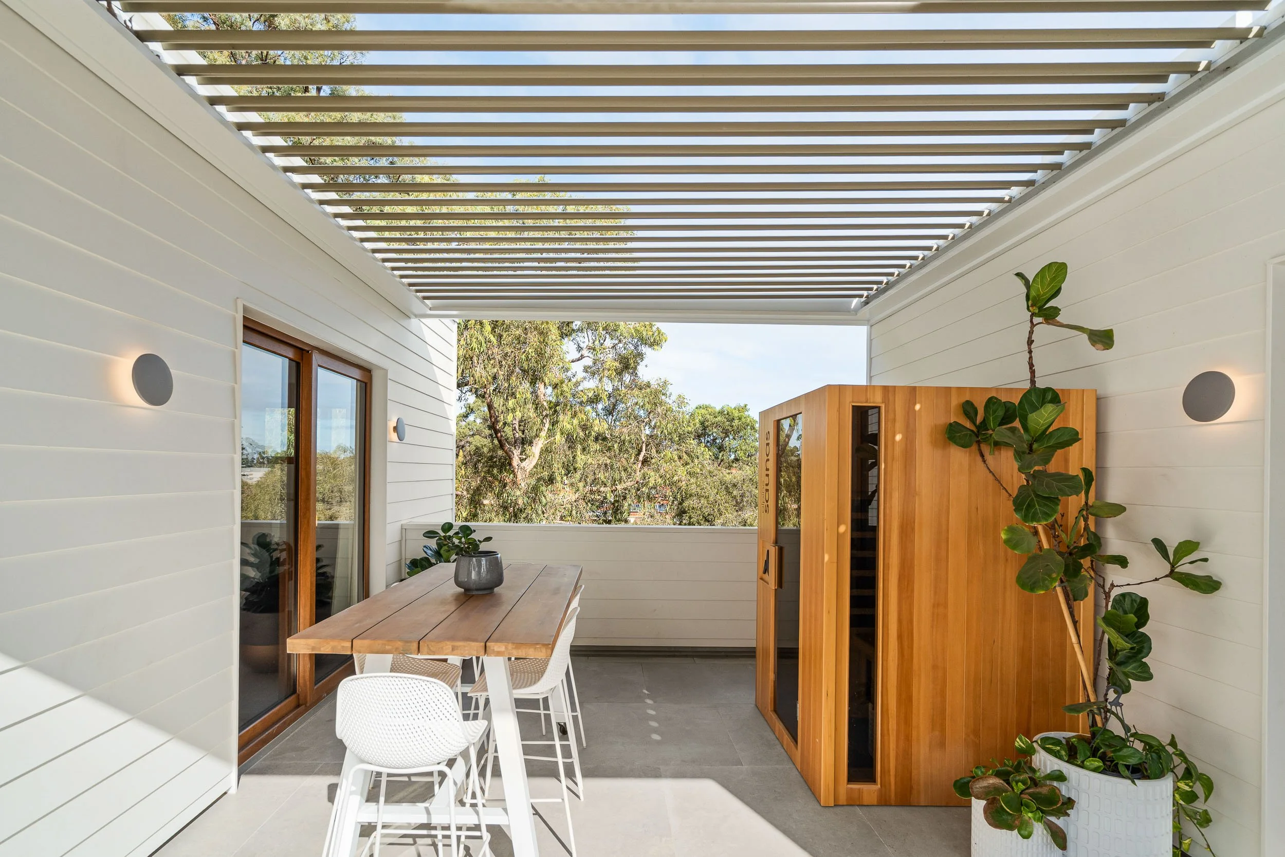 Outdoor terrace with dining furniture beneath a slatted pergola and a timber privacy screen.