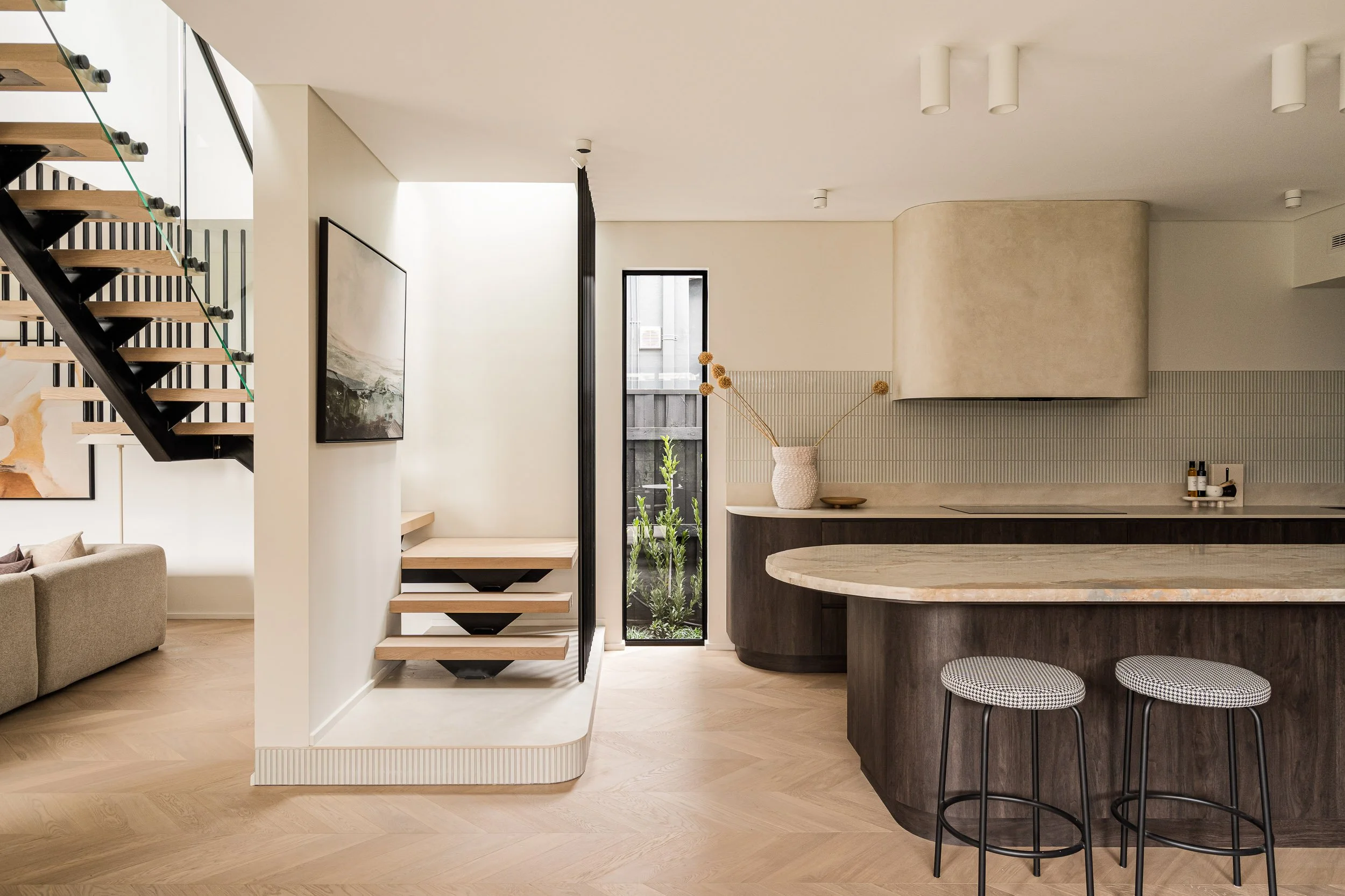 Architectural kitchen with a curved island, pale herringbone flooring and a floating timber staircase.