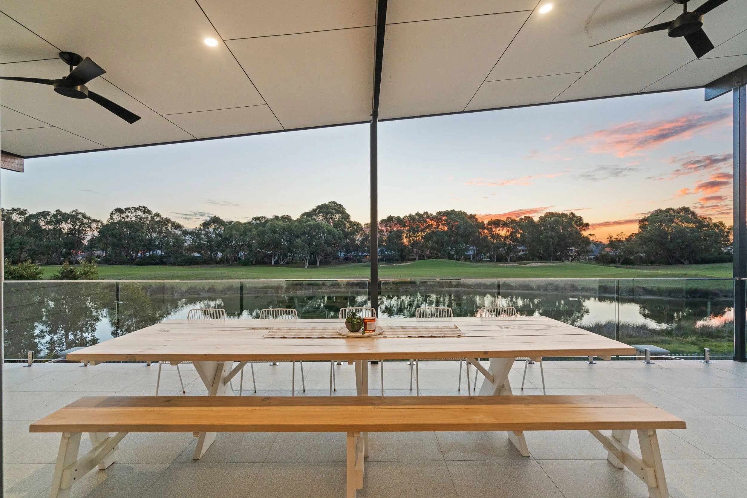 Covered outdoor dining area with glass balustrades overlooking the water at sunset.