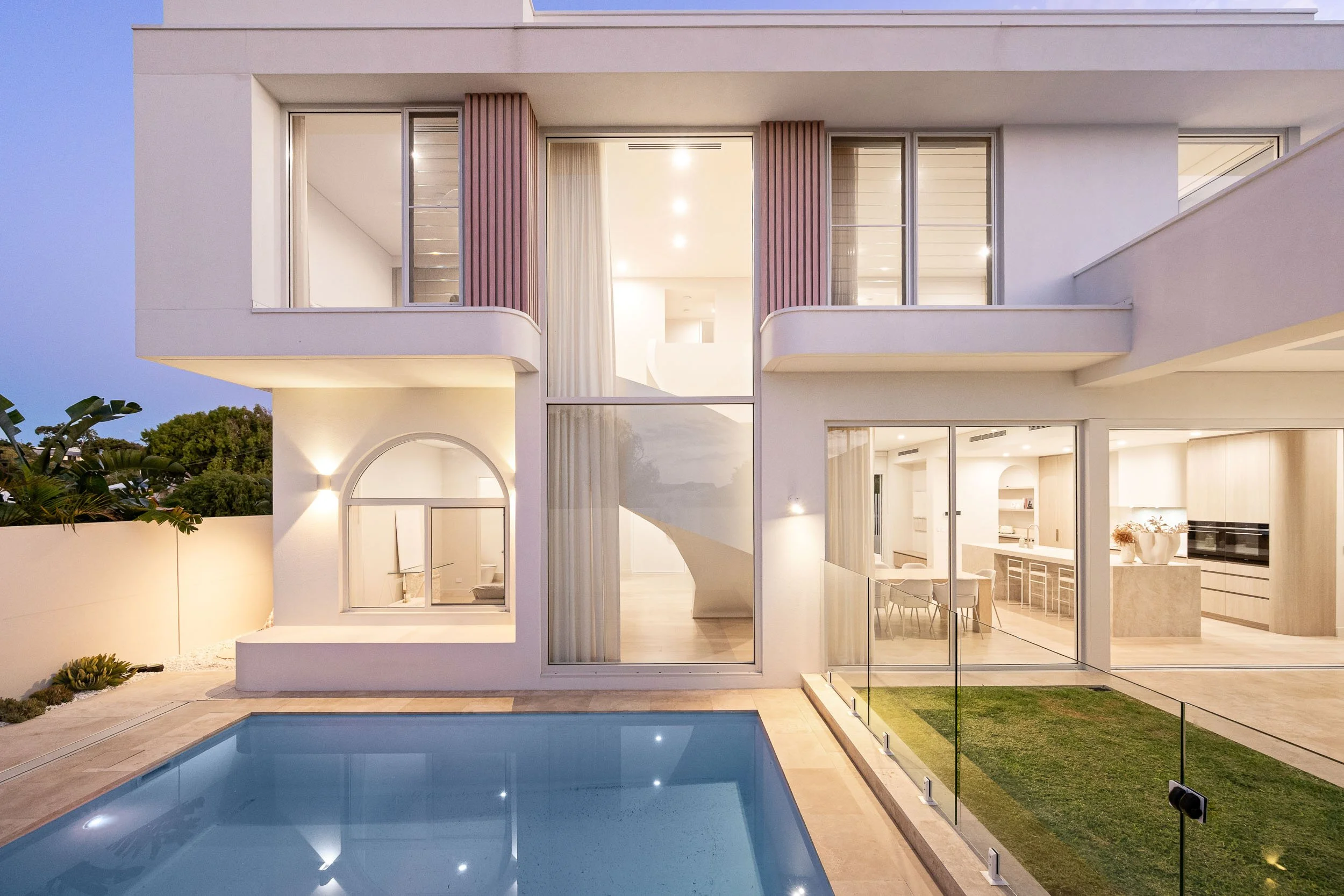 Two-storey contemporary facade at twilight with tall glazing, soft curtains and a pool in the foreground.