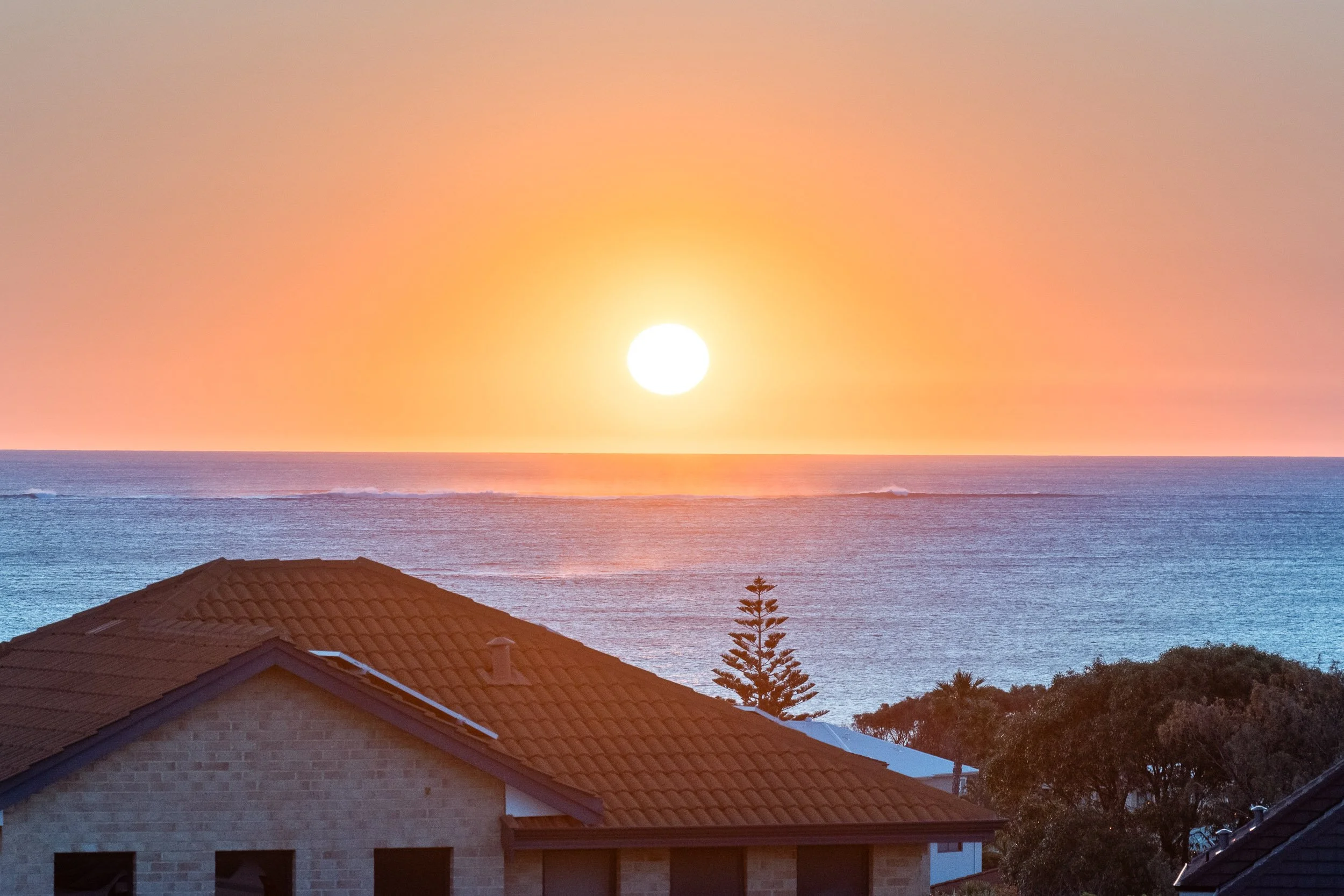 Orange sunset over the ocean viewed above coastal rooftops.
