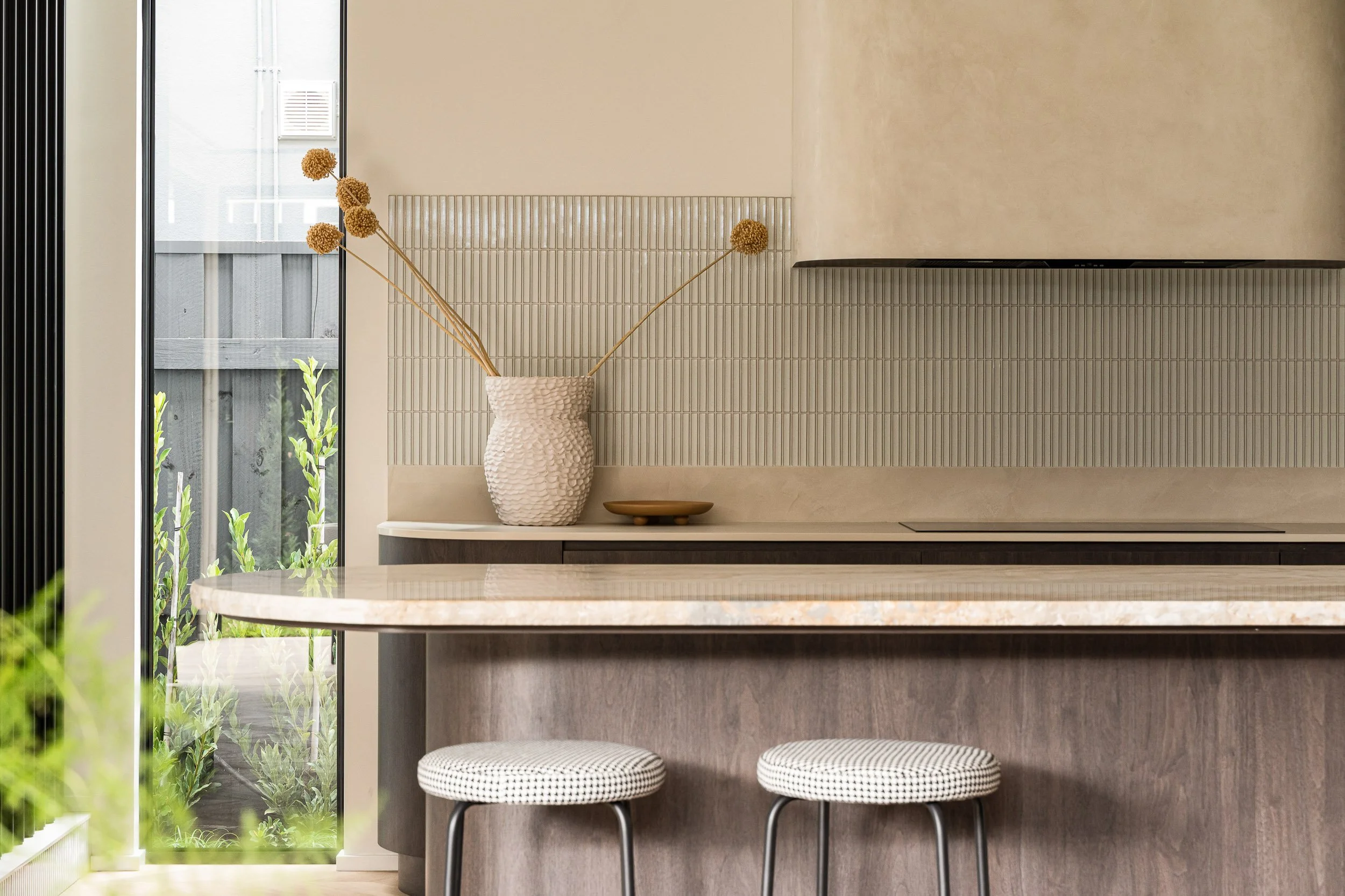Kitchen detail with a stone island, two woven stools, slim tiled splashback and soft natural light.