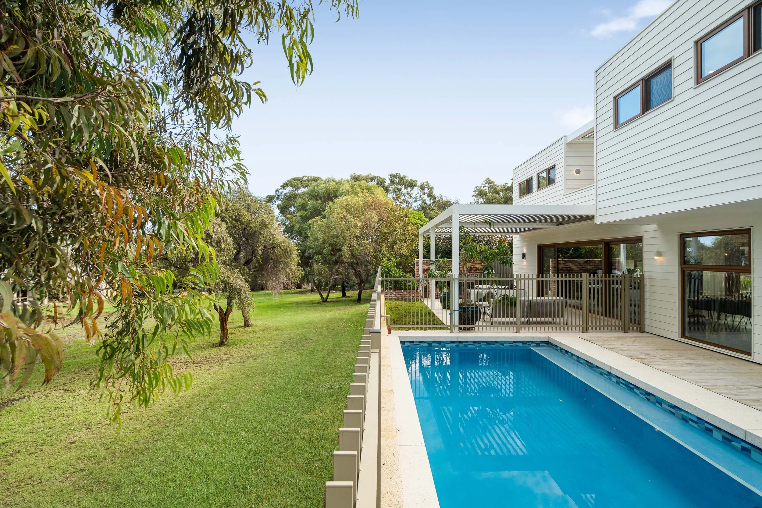 Swimming pool beside a contemporary white-clad home and a tree-lined lawn.