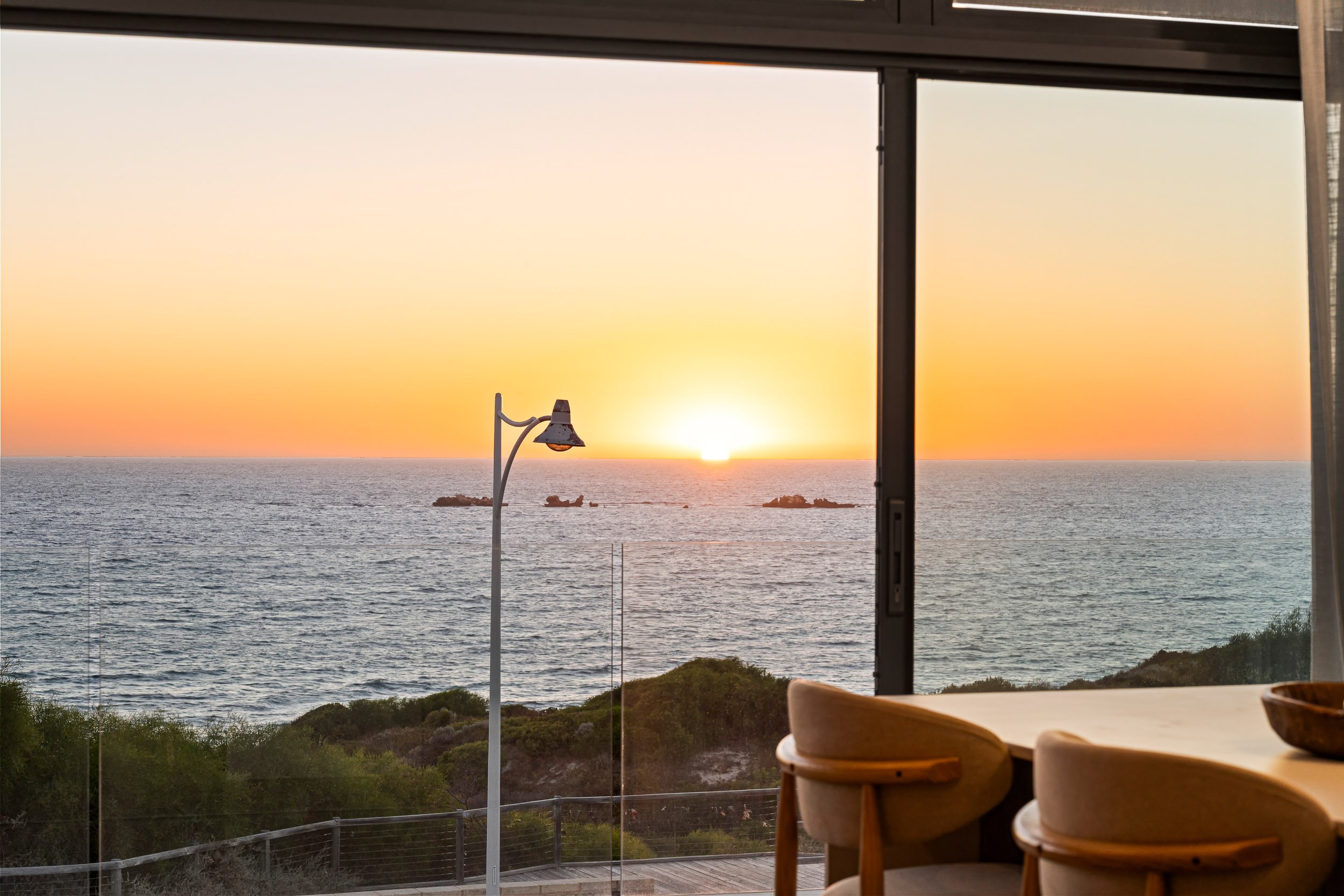 Ocean sunset viewed through a full-height window beside a small table and chair.