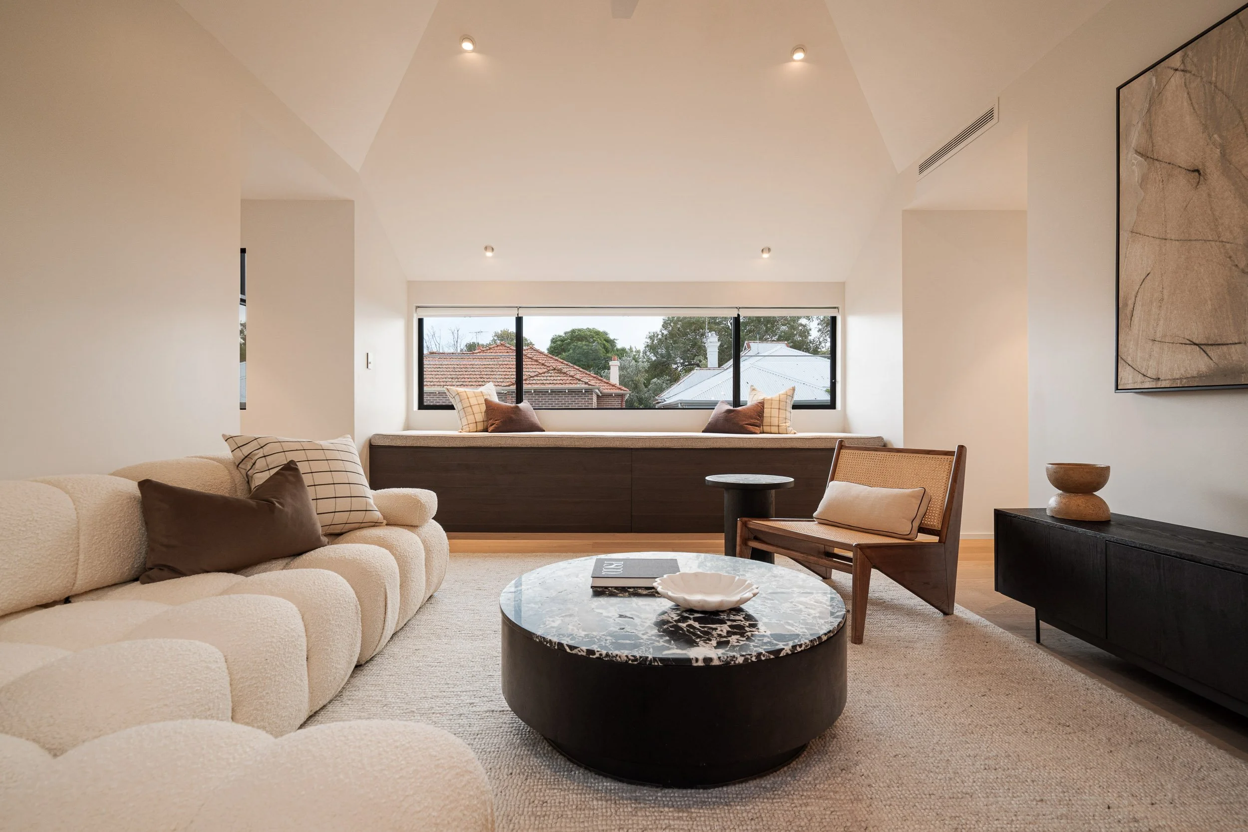 Living room with a curved sofa, round dark coffee table and timber joinery beneath a wide window.