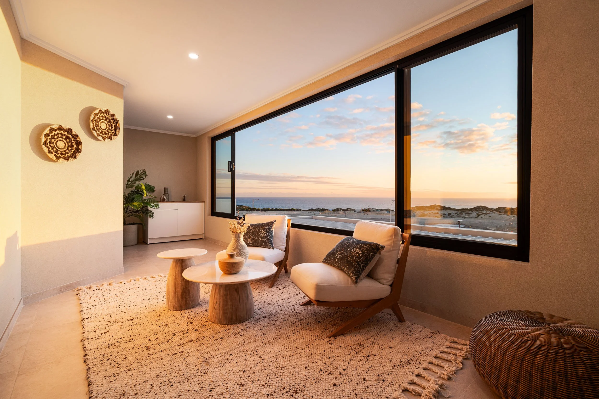 Sunlit sitting area with lounge chairs and broad windows overlooking the coast at sunset.