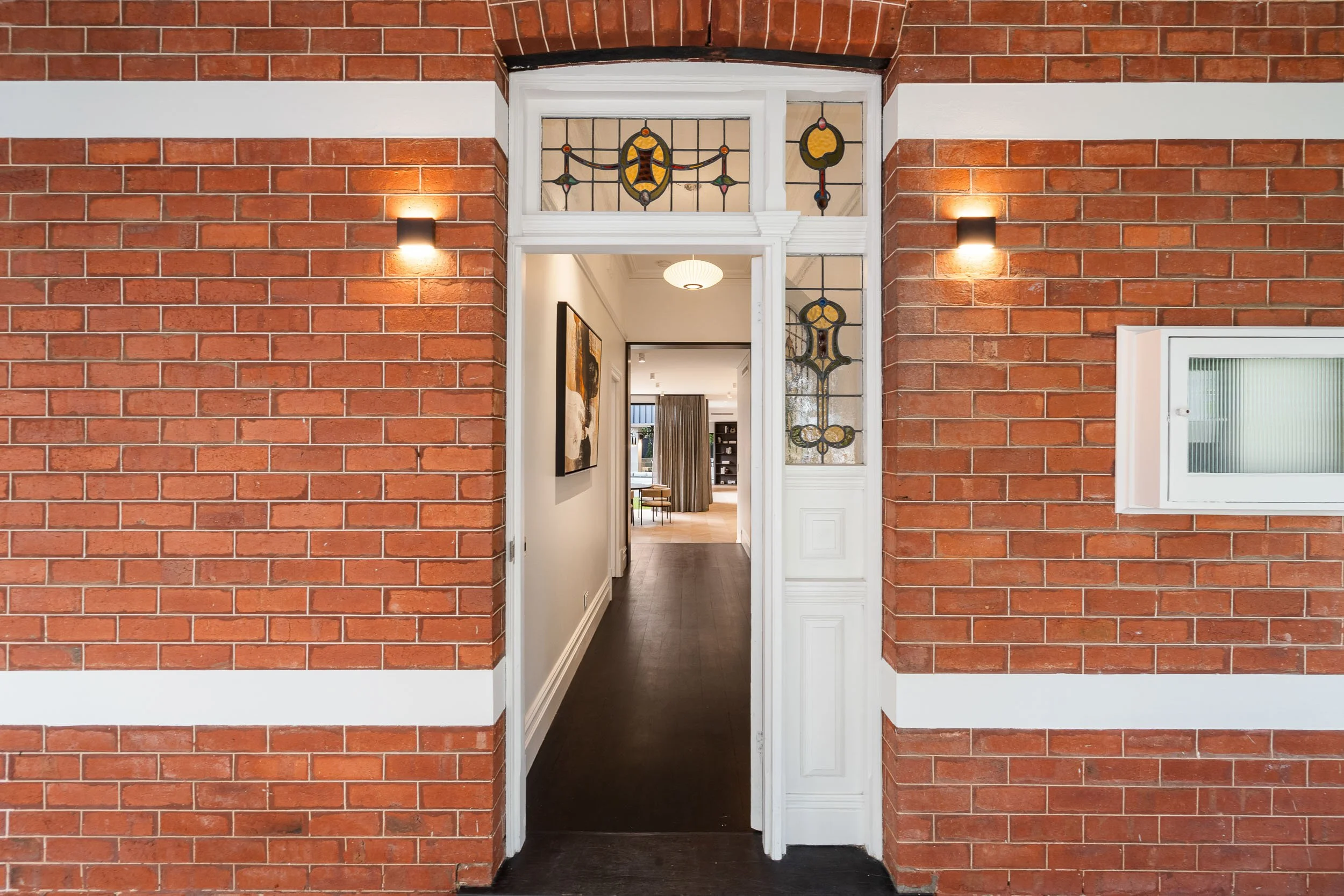 Red-brick home entrance with decorative leadlight glass, a white timber door and a view along the hallway.