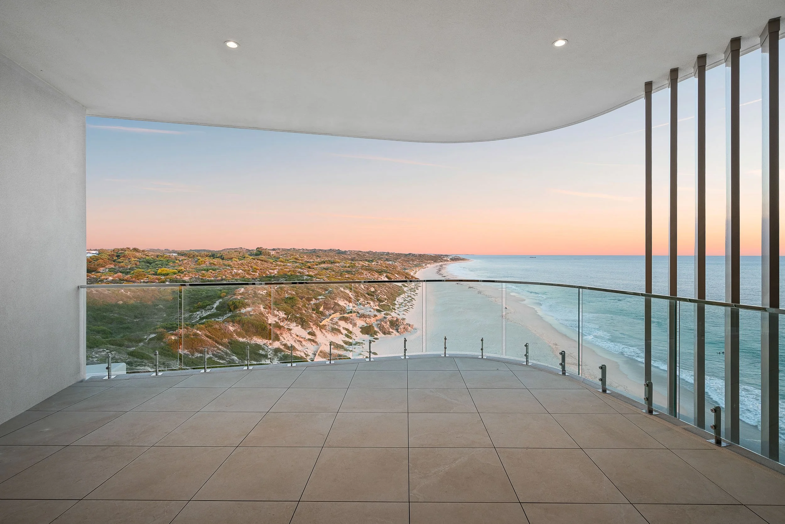 Covered coastal balcony with glass balustrades and a sweeping beach view at sunset.