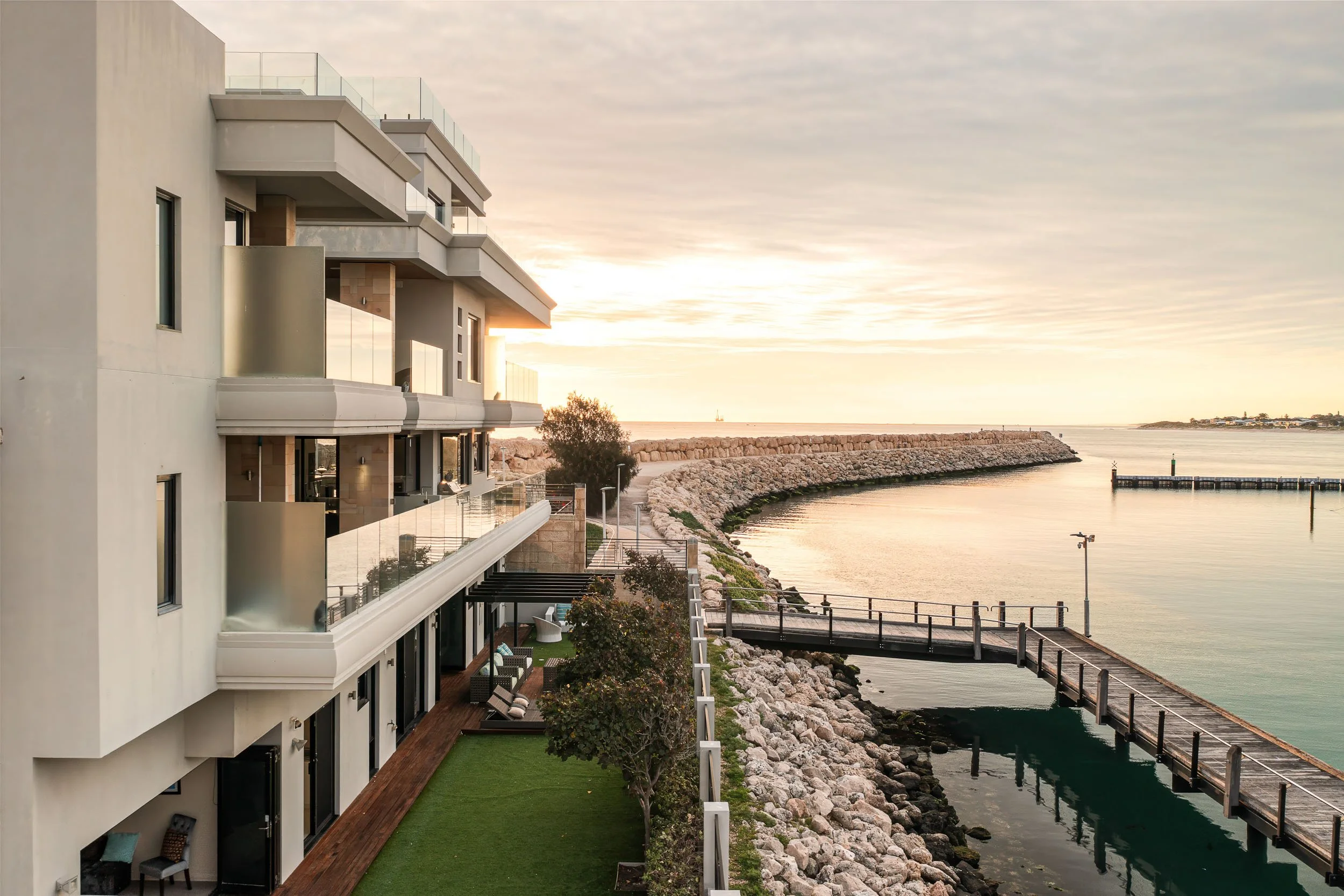 Contemporary waterfront building with balconies overlooking a sheltered harbour, timber boardwalk and stone breakwater at sunset.
