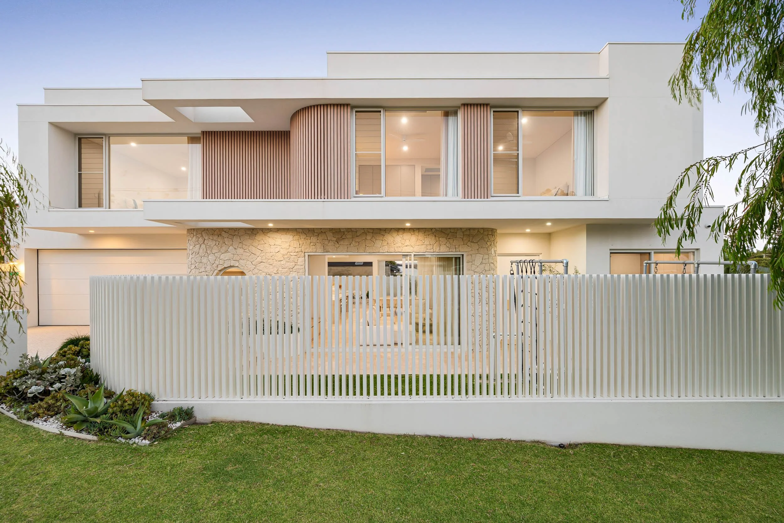 Contemporary two-storey home at twilight with a curved upper facade, white slatted fence and landscaped lawn.